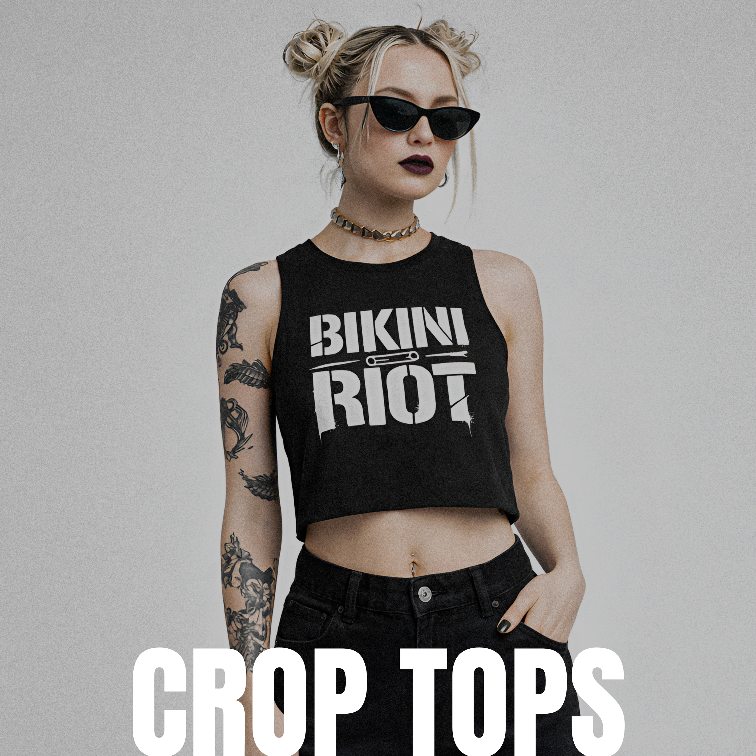 A young woman with tattoos, wearing black sunglasses, a black sleeveless crop top with 'Bikini Riot' print, and black pants. She has blonde hair styled in space buns and wears dark lipstick and accessories, posing against a plain background.