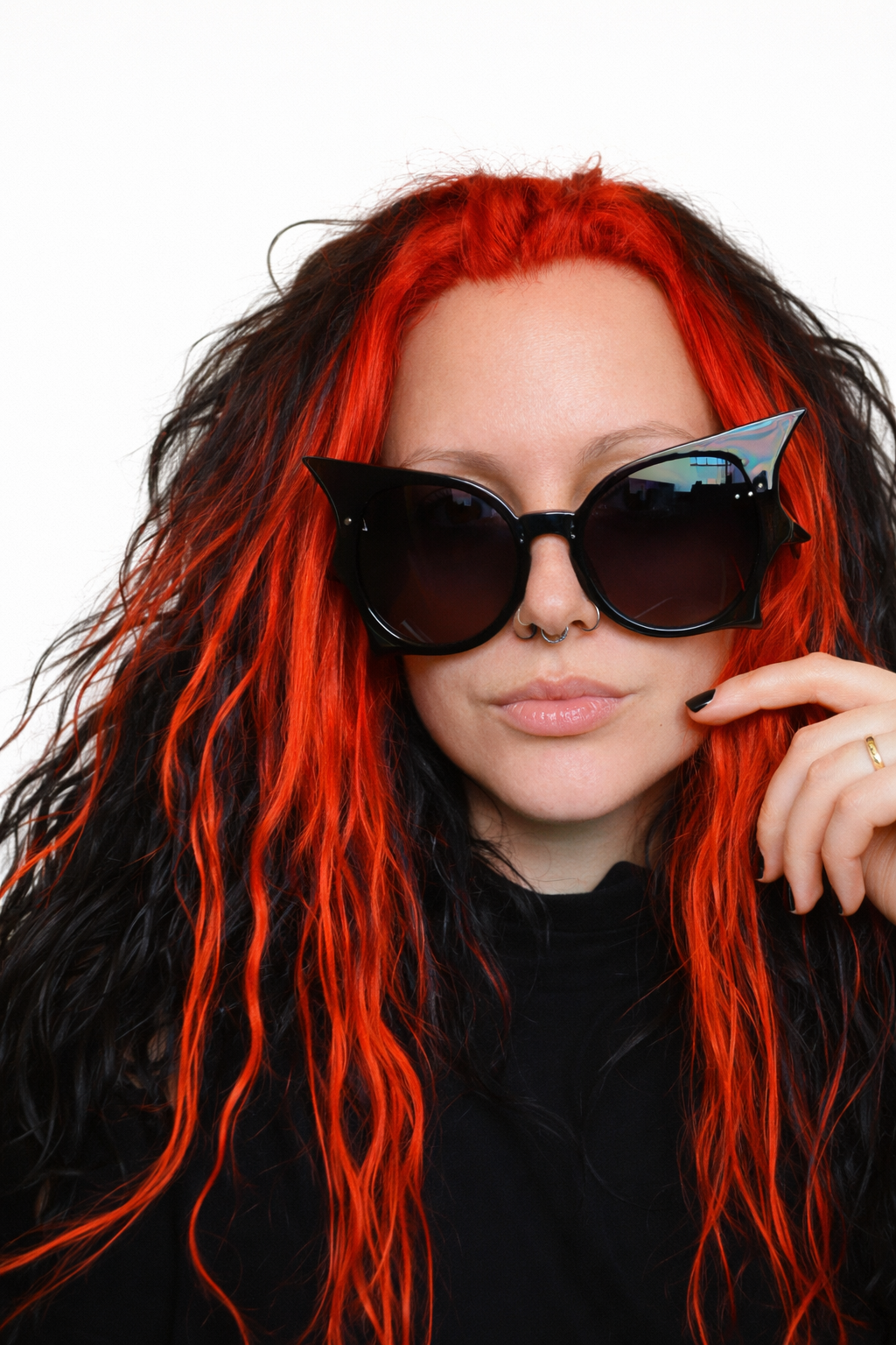 A woman with long red and black hair wearing large black sunglasses, a septum piercing, and a black top.