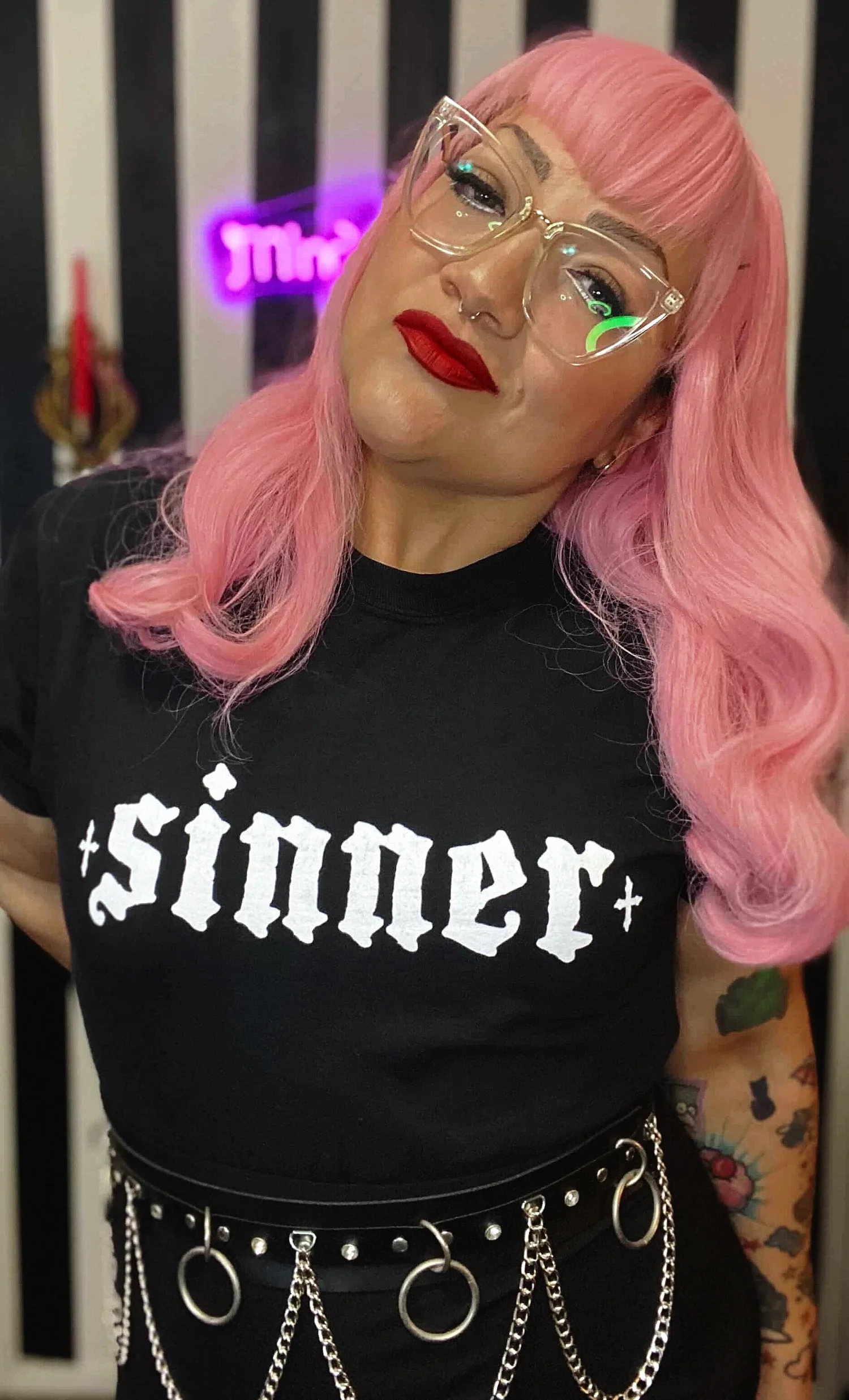 A woman with pink hair, wearing clear glasses, red lipstick, a black shirt with white Gothic-style text, and a black belt with metal chains and rings, posing indoors with a neon sign in the background.