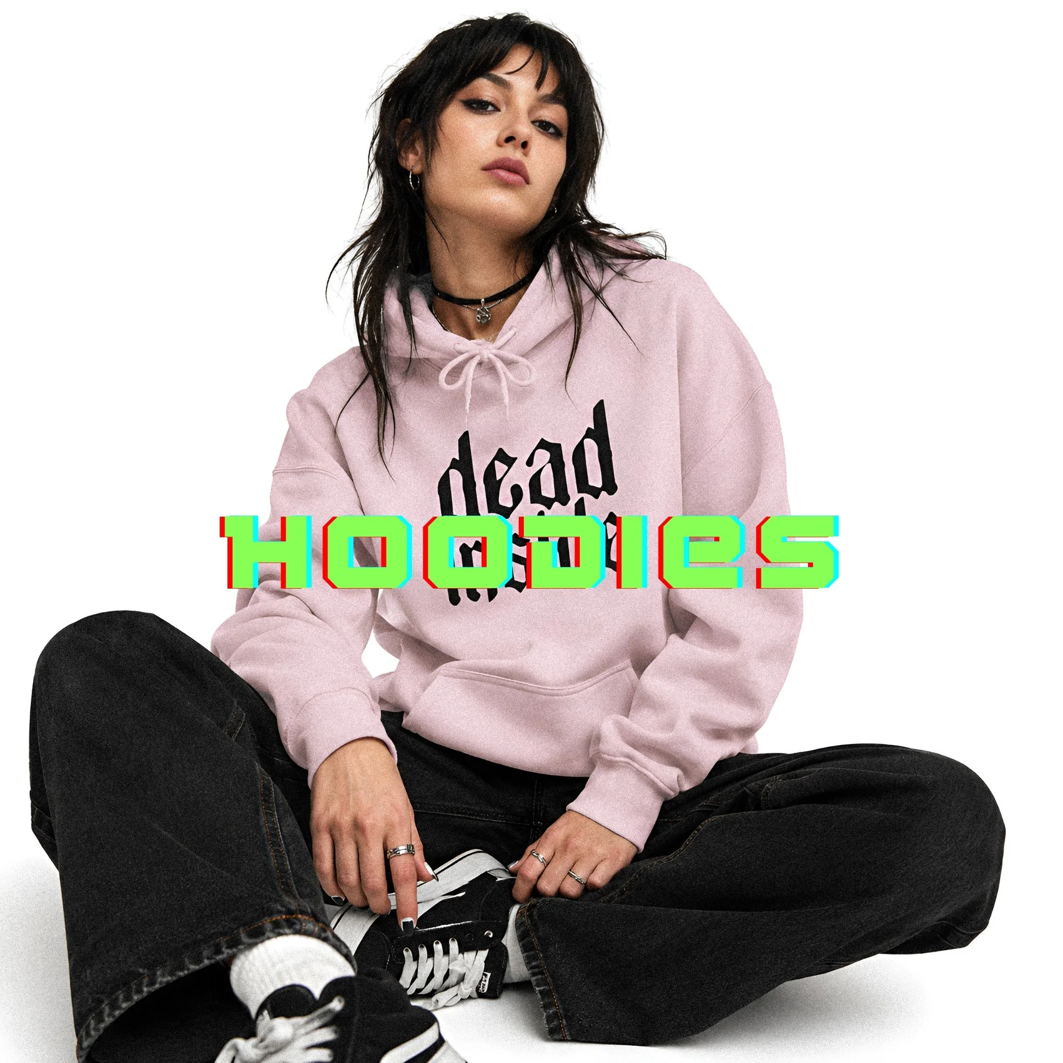 A young woman with dark hair and makeup, wearing earrings and layered necklaces, sitting against a white background, dressed in a pink hoodie with black text, black pants, and black and white sneakers.