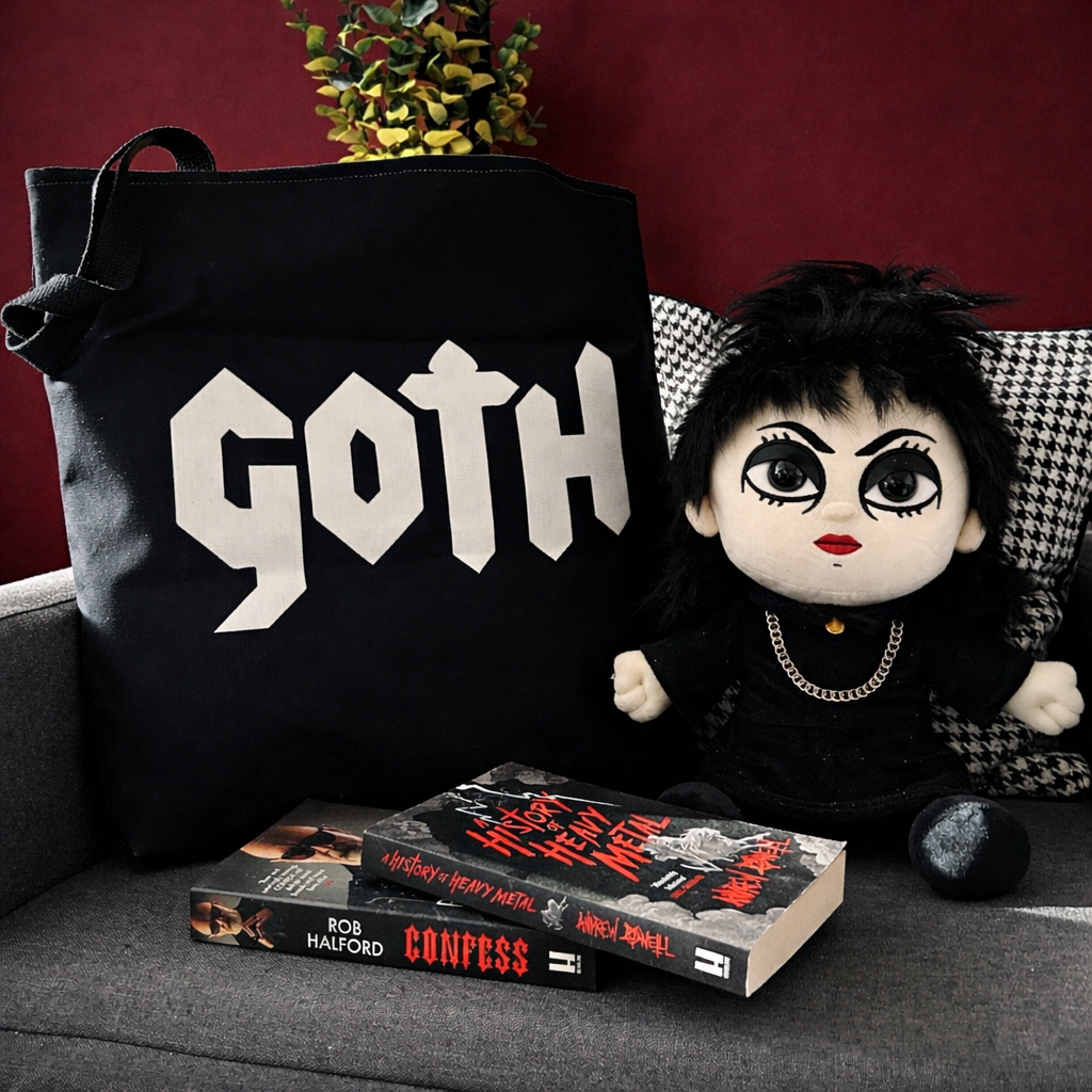 A black tote bag with the word 'Goth' in white letters, a doll with black hair, dark eye makeup, red lipstick, wearing black clothing with chains, sitting on a couch with houndstooth and solid pillows, and three heavy metal books by Rob Halford and Andrew Darrell on the couch.