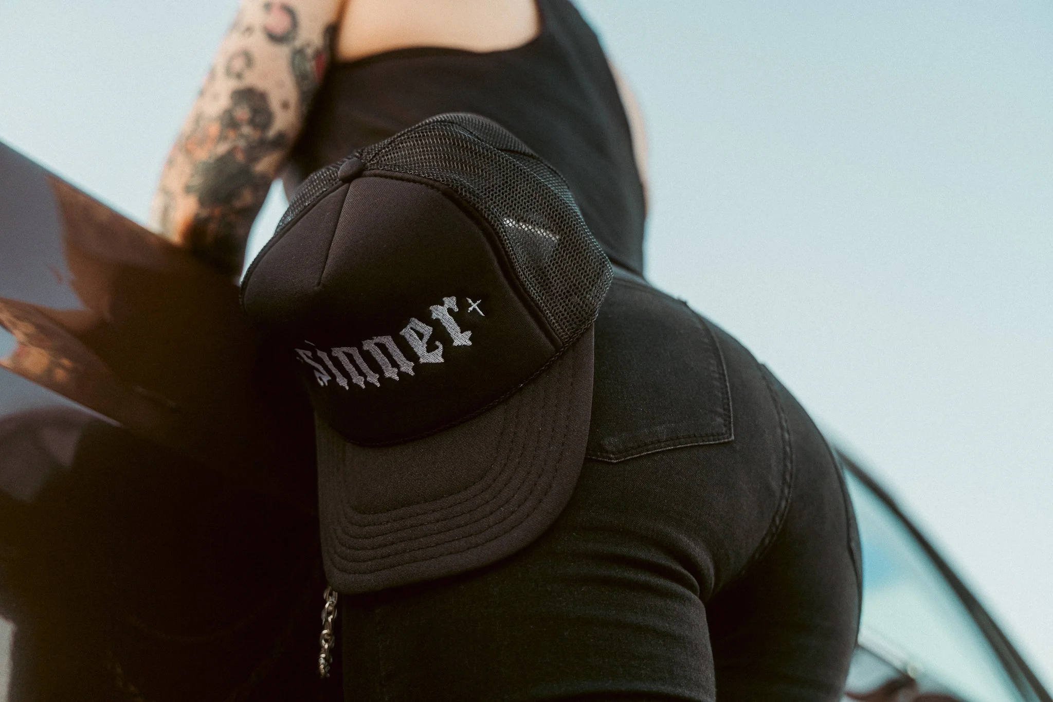 Close-up of a person's arm with tattoos, wearing a black baseball cap with white text and a black sleeveless top.