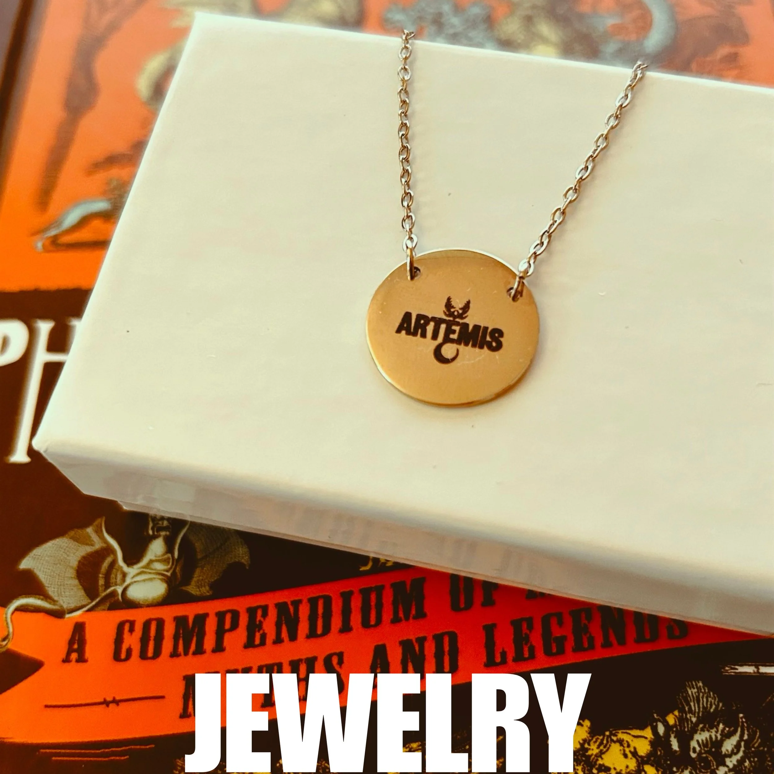 Gold necklace with a circular pendant engraved with the word 'ARTEMIS' and a small winged symbol above it, resting on a white jewelry box with books in the background.