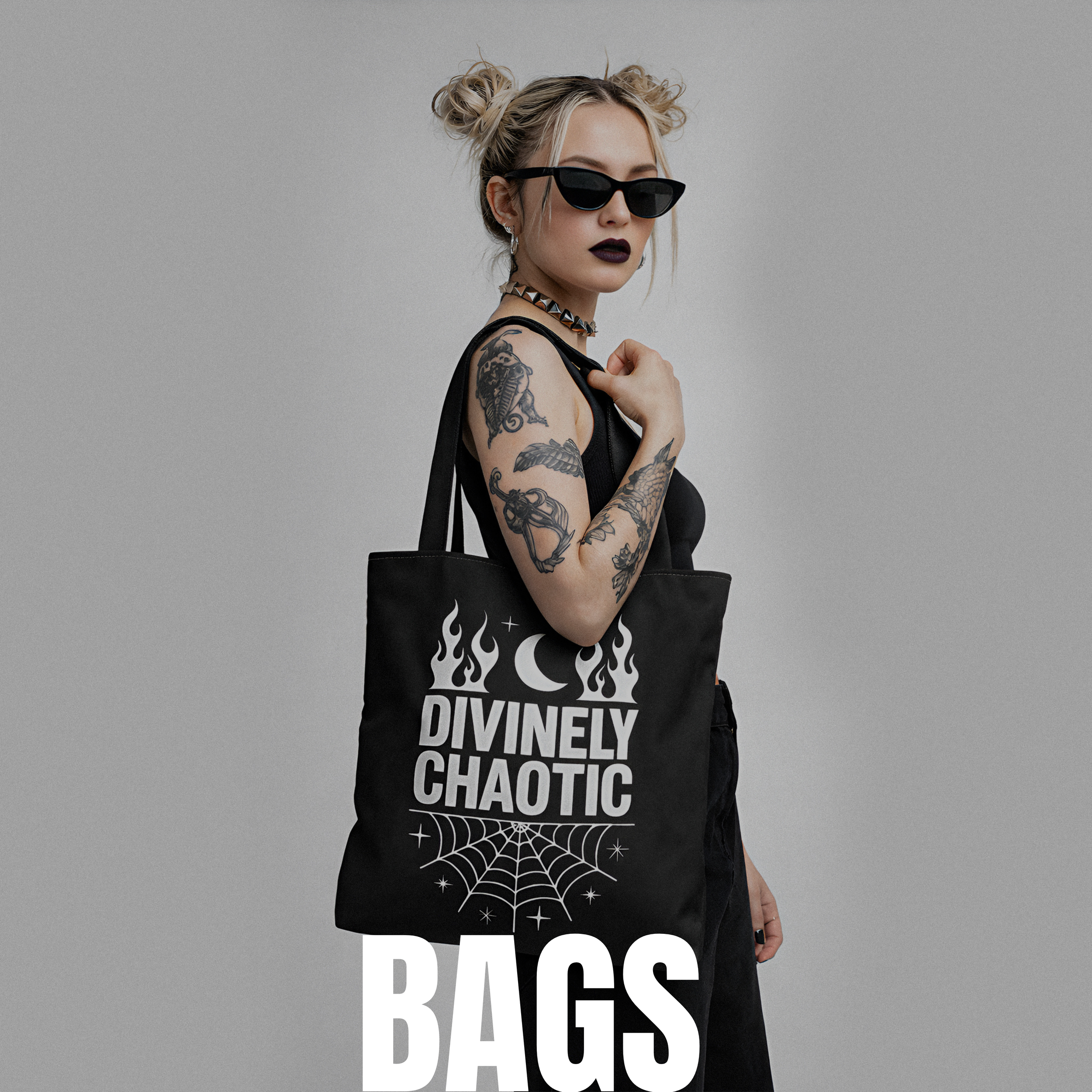 A woman with blonde hair styled in space buns, wearing black sunglasses, dark lipstick, and a black sleeveless top, carrying a black tote bag with white text and graphic design. The tote bag reads "Divinely Chaotic" with a moon, flames, spider web, and stars. The image has a plain gray background.