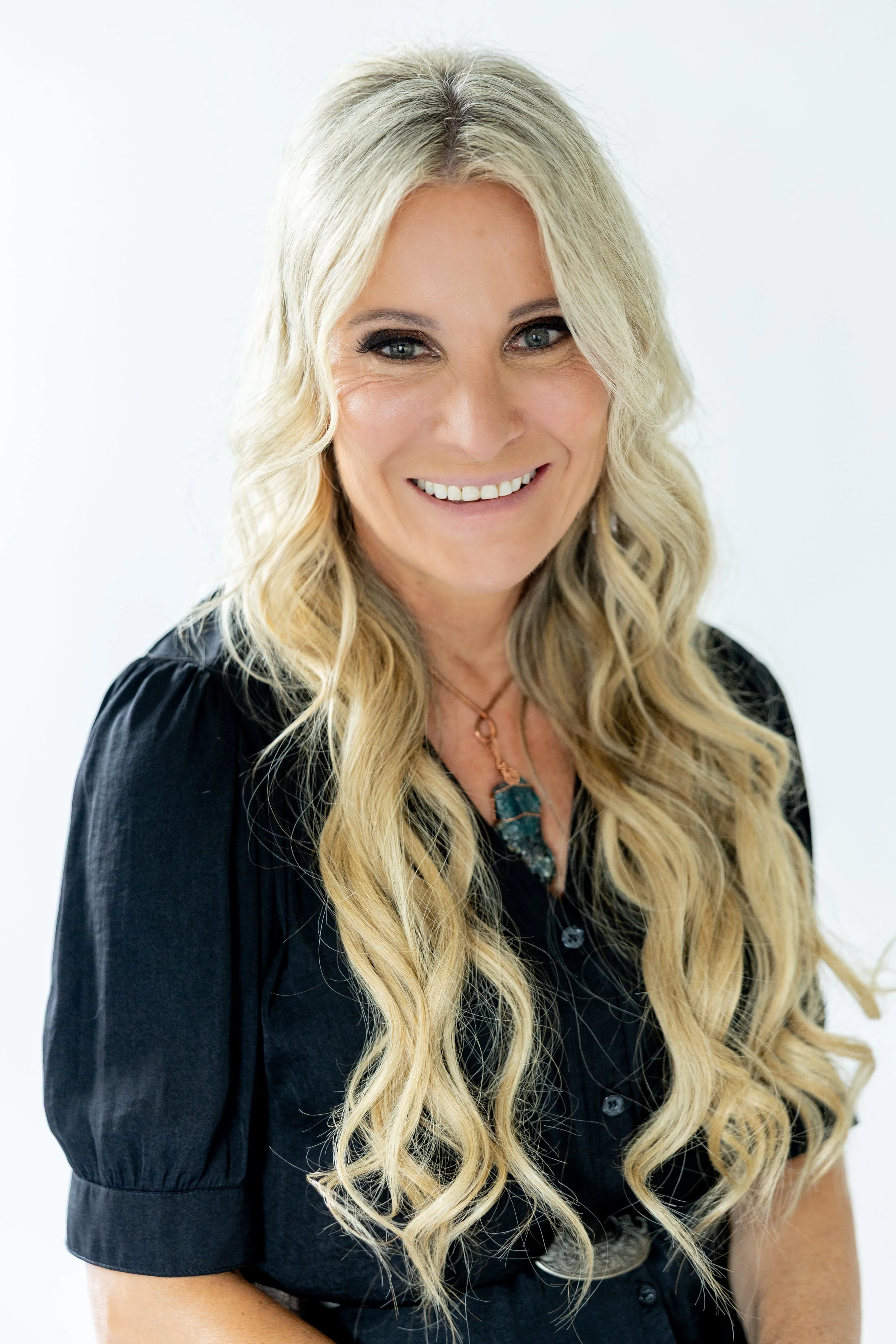 Deborah Mottola , Crystal Expert, Wisdom Teacher, A middle-aged woman with long, wavy blonde hair smiling at the camera, wearing a black top and a crystal necklace with a large, dark-colored pendant.