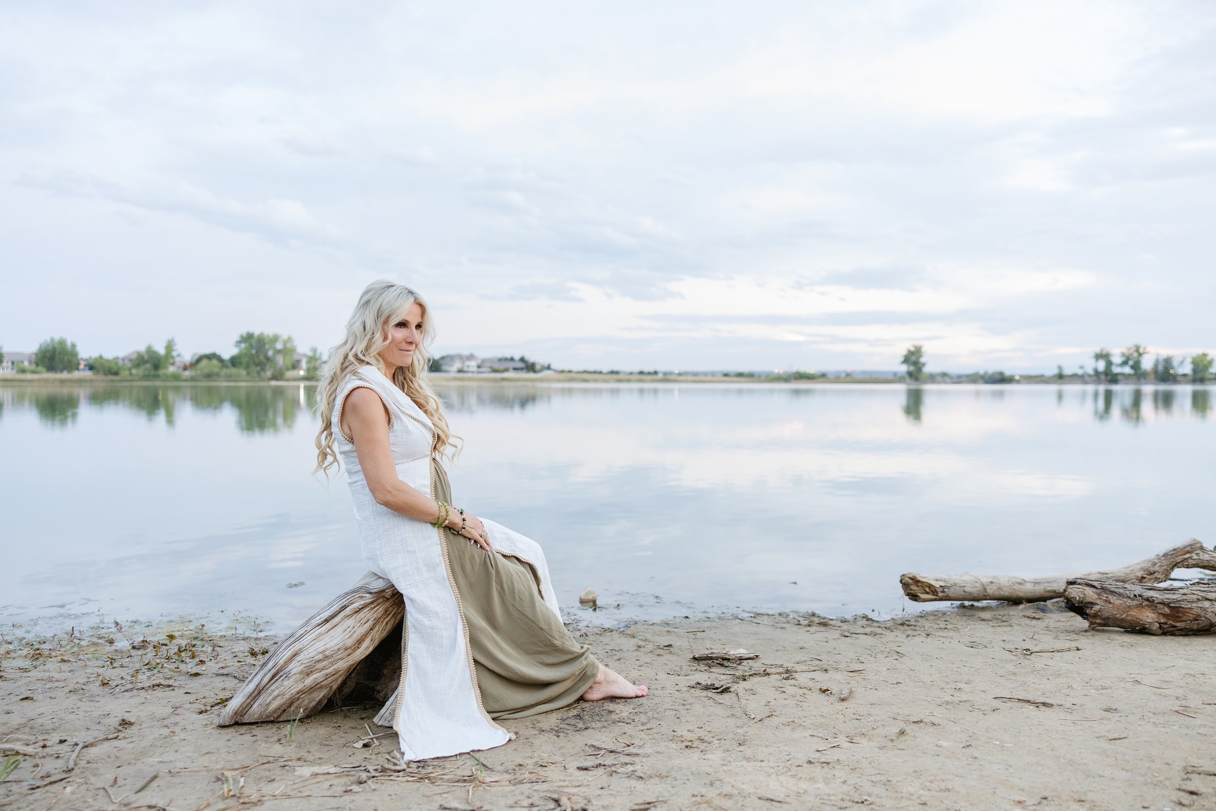 Energy Healing, Ascension A woman with long blonde hair sitting on a log by a calm lake, with a desert landscape and cloudy sky in the background.