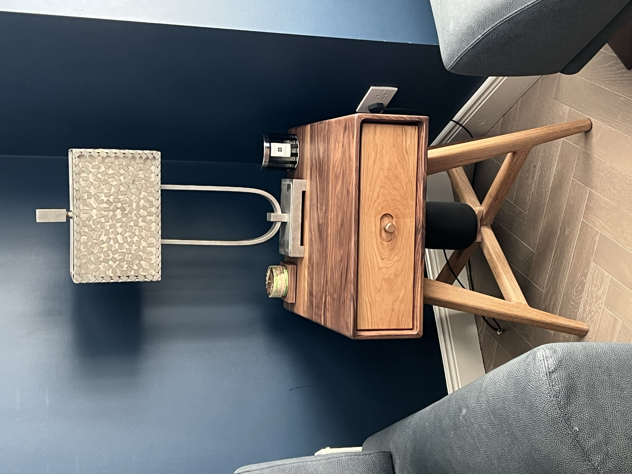 A wooden desk with a drawer, a small candle, and a metallic lamp on a navy blue wall background.