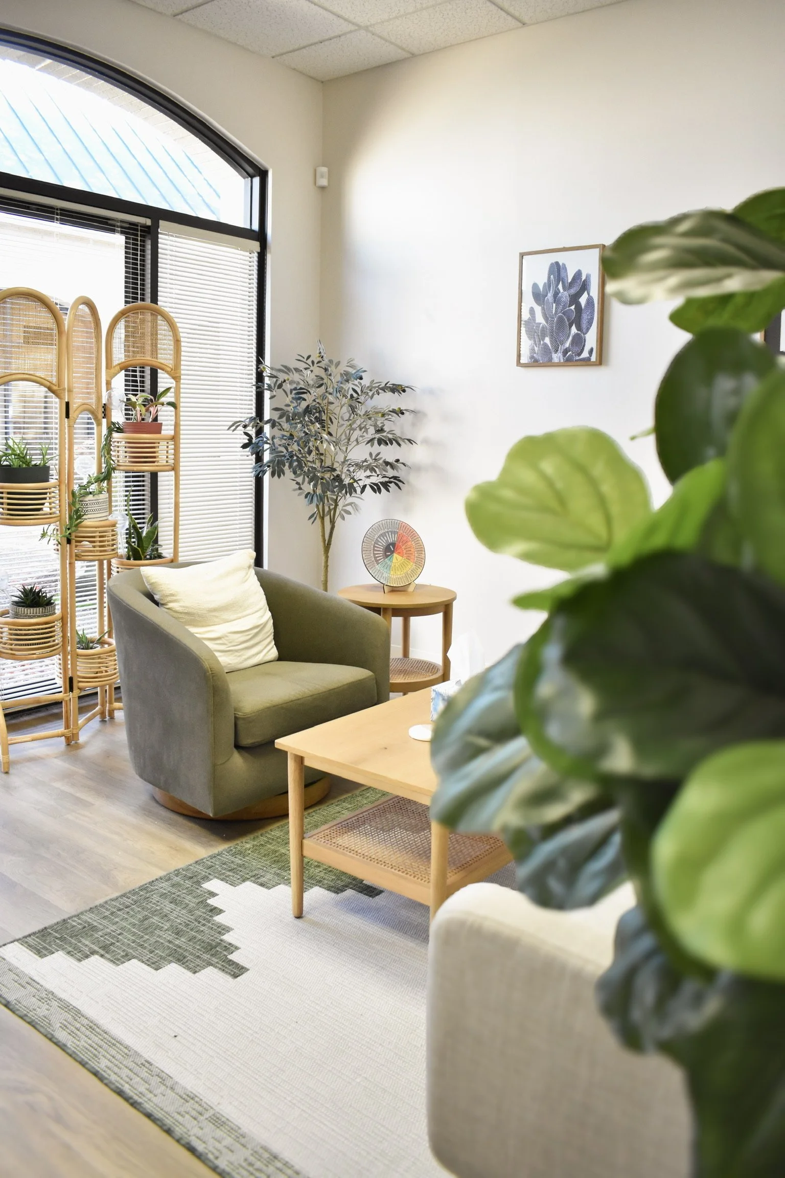 A cozy indoor space with a gray armchair, a small wooden side table, and green plants. There's a framed picture of cacti on the wall. A woven room divider holds small potted plants near a large window with blinds.