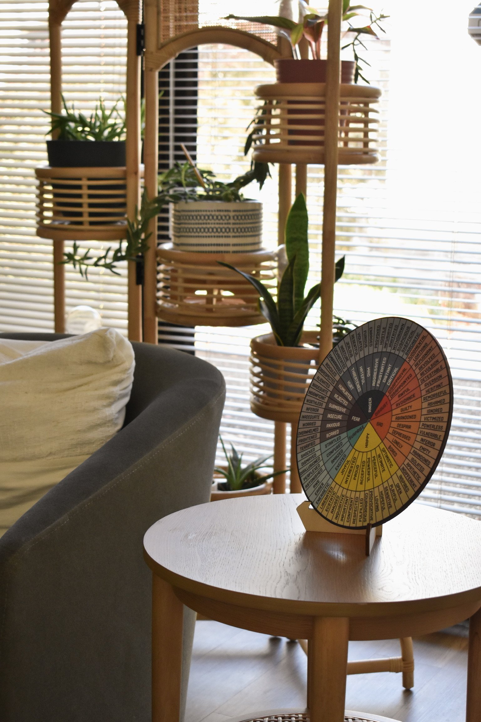 Interior view of a living room with plants on shelving unit, window blinds in the background, part of a gray sofa with a white pillow, and a round wooden table holding a feelings wheel.