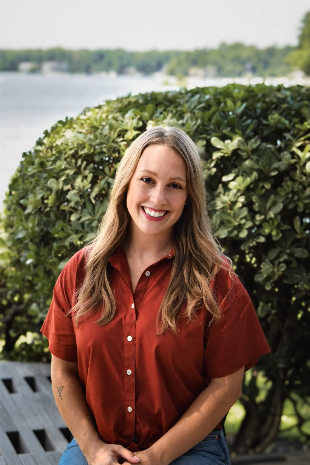A smiling woman with long light brown hair, wearing a short-sleeved rust-colored button-up shirt, sitting outdoors with lush green foliage and a body of water in the background.