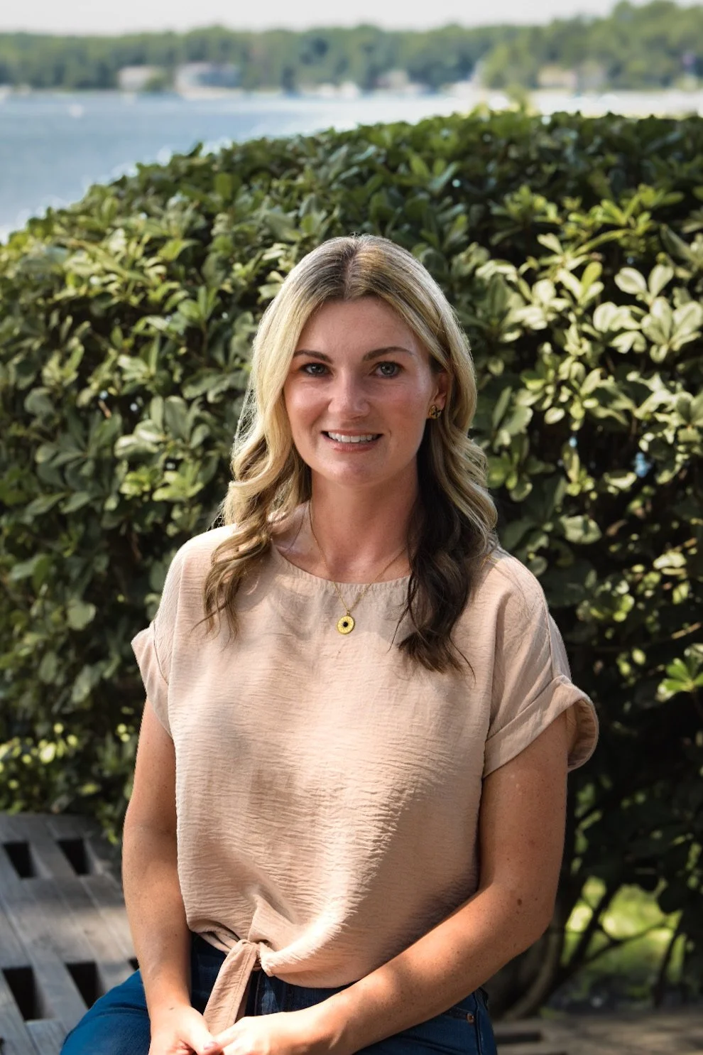 A woman with blonde wavy hair, wearing a light beige top and blue jeans, sitting outdoors near green shrubs and a body of water in the background.