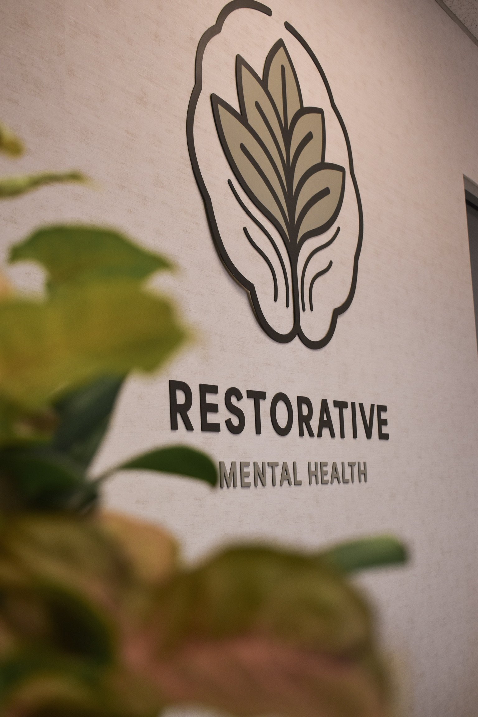 Sign for Restorative Mental Health services with logo and blurred plants in the foreground.