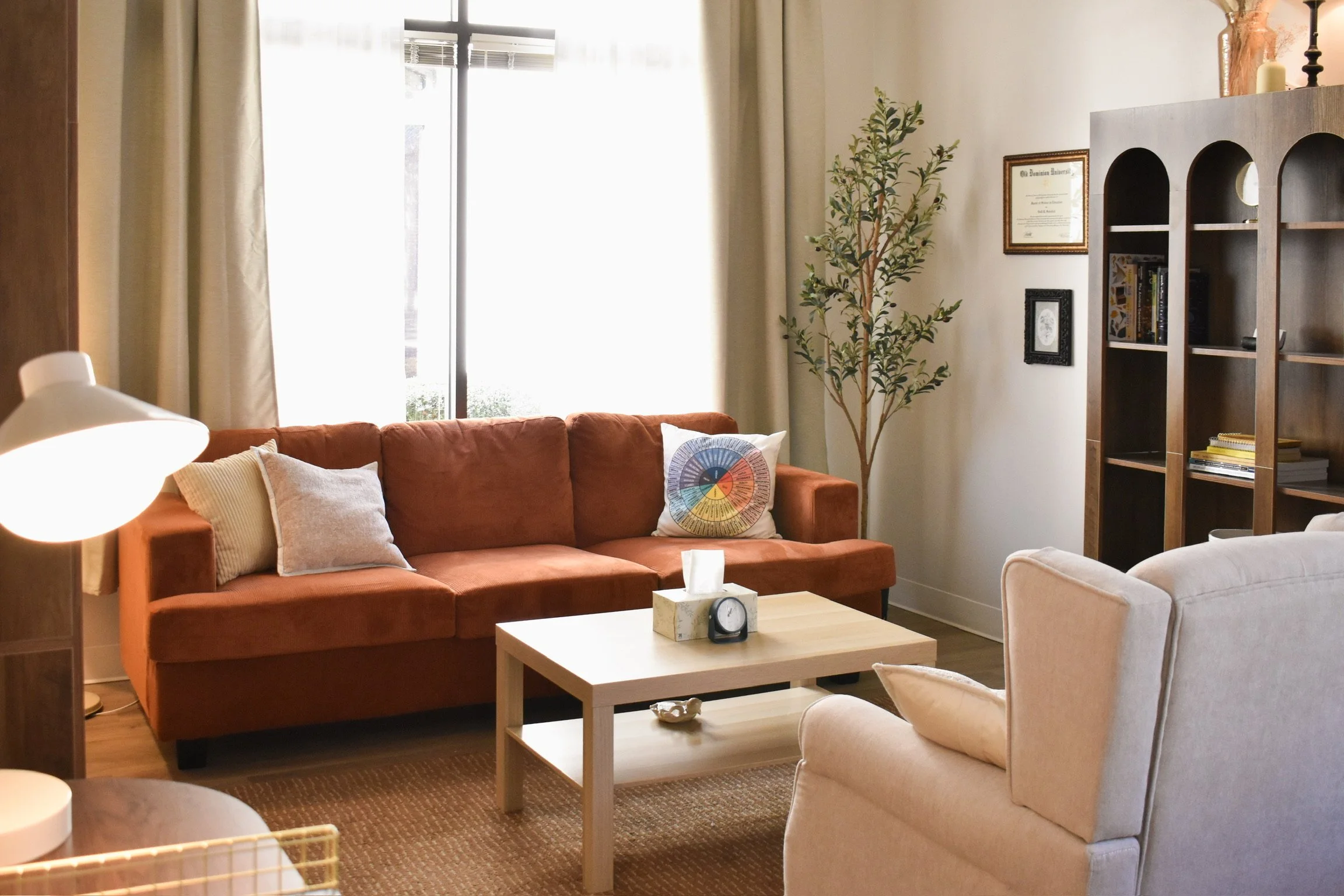 Sophisticated office with an orange sofa, a white armchair, a wooden coffee table, a tall potted plant, a wooden bookshelf, and framed artwork on the wall. Bright sunlight coming through a large window.