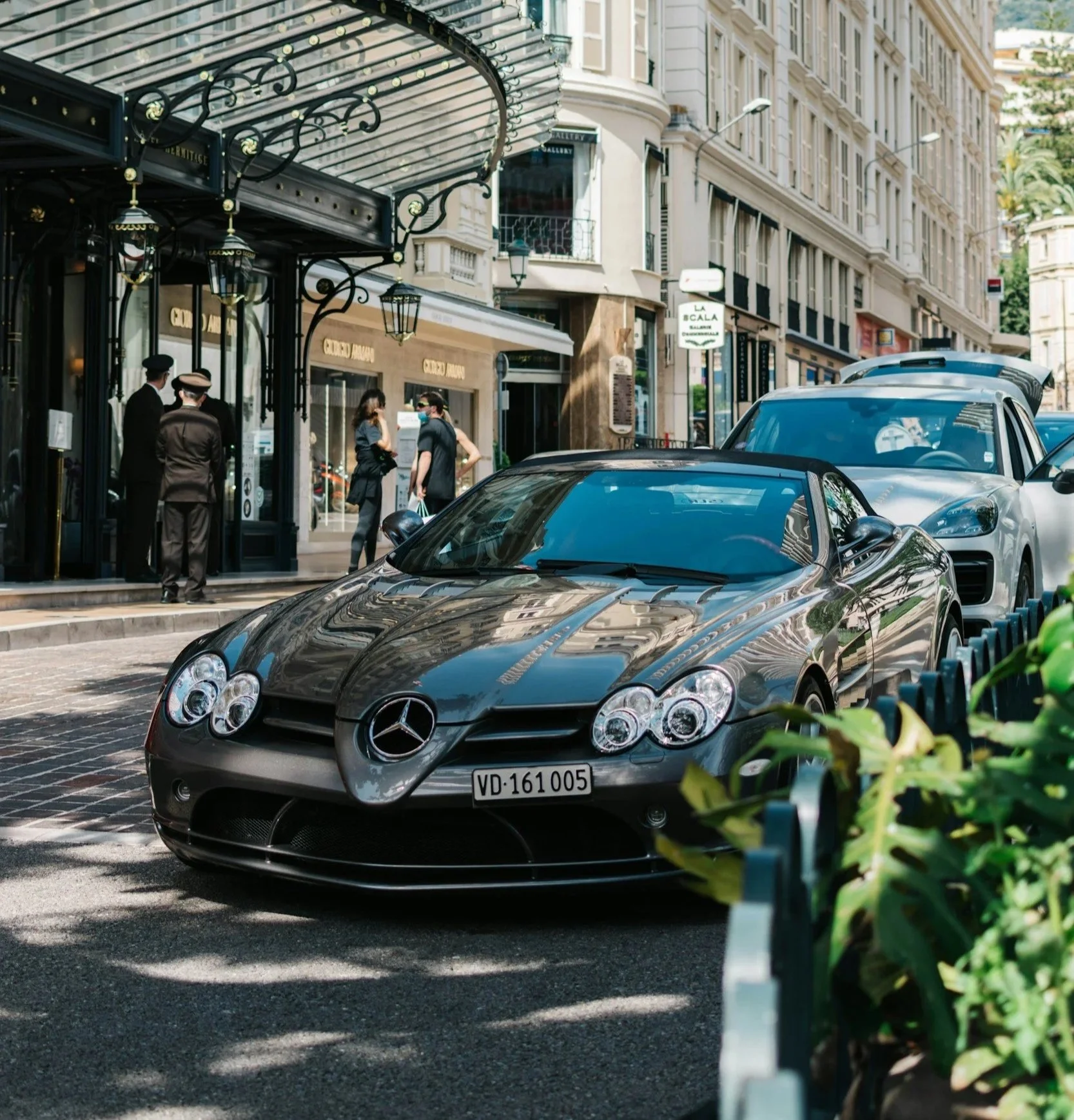 Schwarzer Mercedes-Benz SLS AMG Sportwagen parkend auf einer Stadtstraße mit mehreren parkenden Autos, umgeben von historischen Gebäuden und Fußgängern, einige tragen Masken.