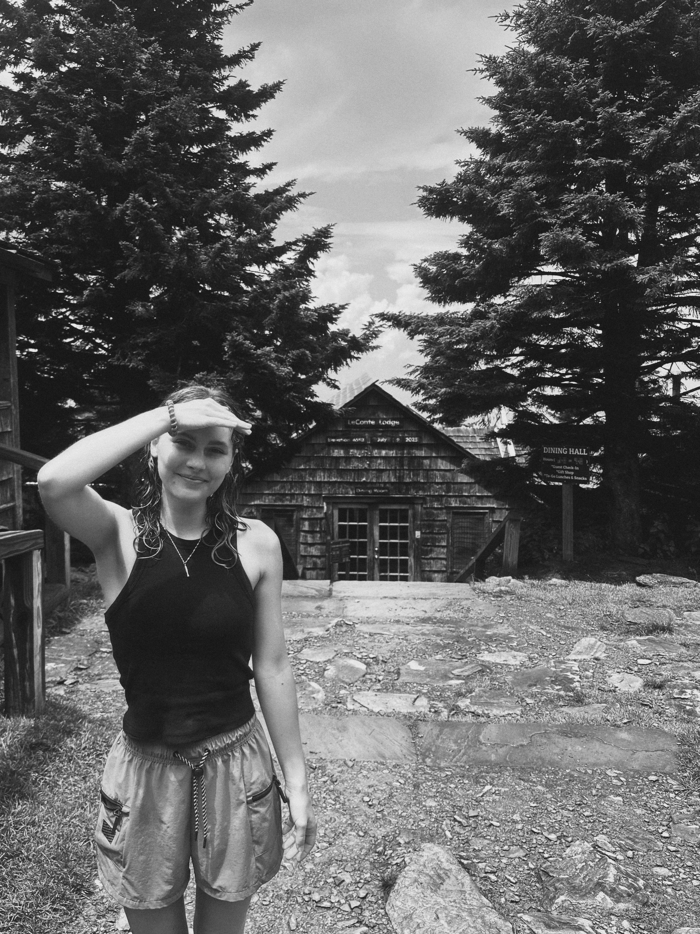 Haley stands beside the dining hall cabin at the summit of Gatlinburg's Mount Leconte, shading her face from the sun with her hand. Her hair is wet because it rained on the hike up the mountain. The cabin is surrounded by pine trees.