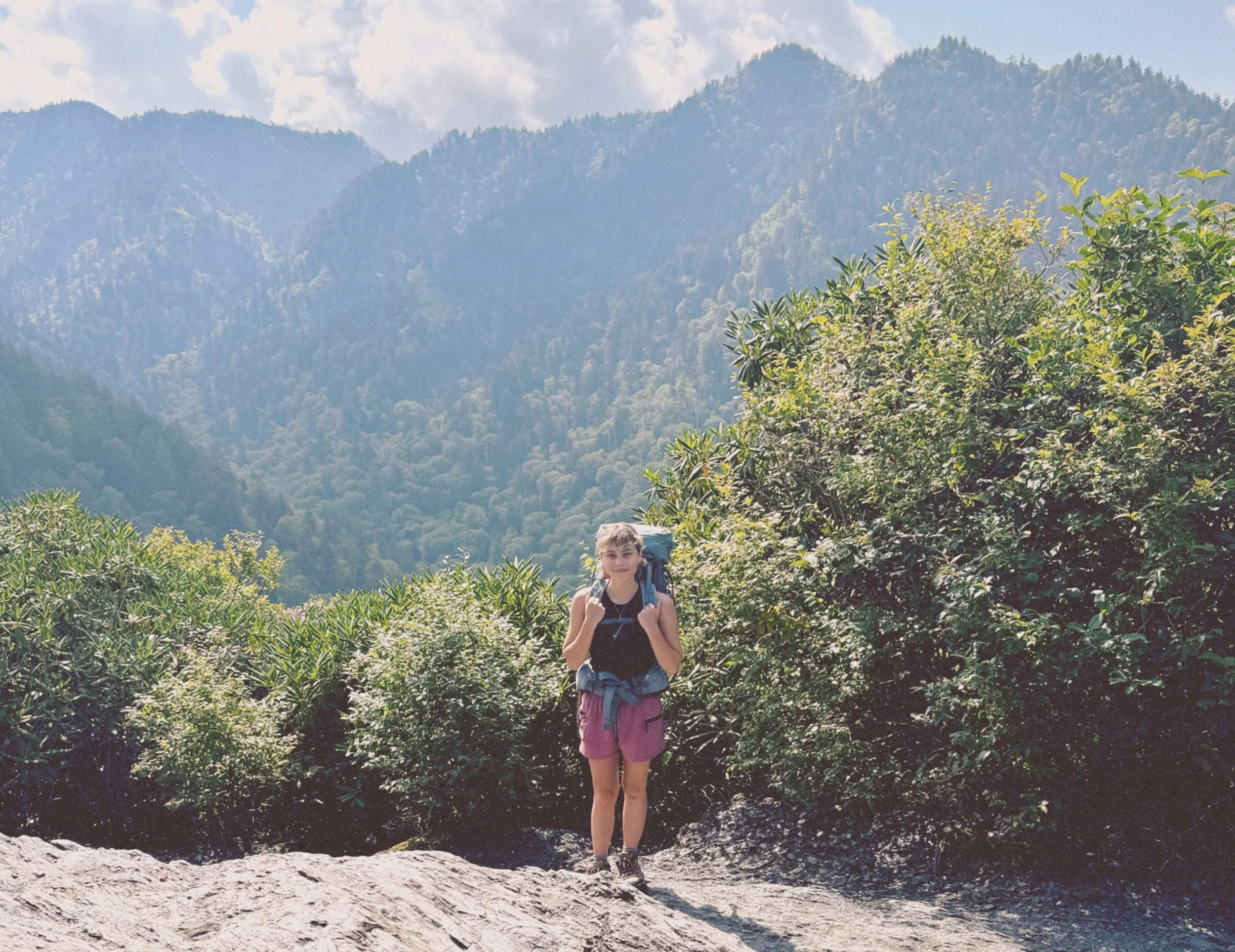 Haley stands against a mountainous backdrop with her blue hiking backpack. She is hiking mount Leconte.