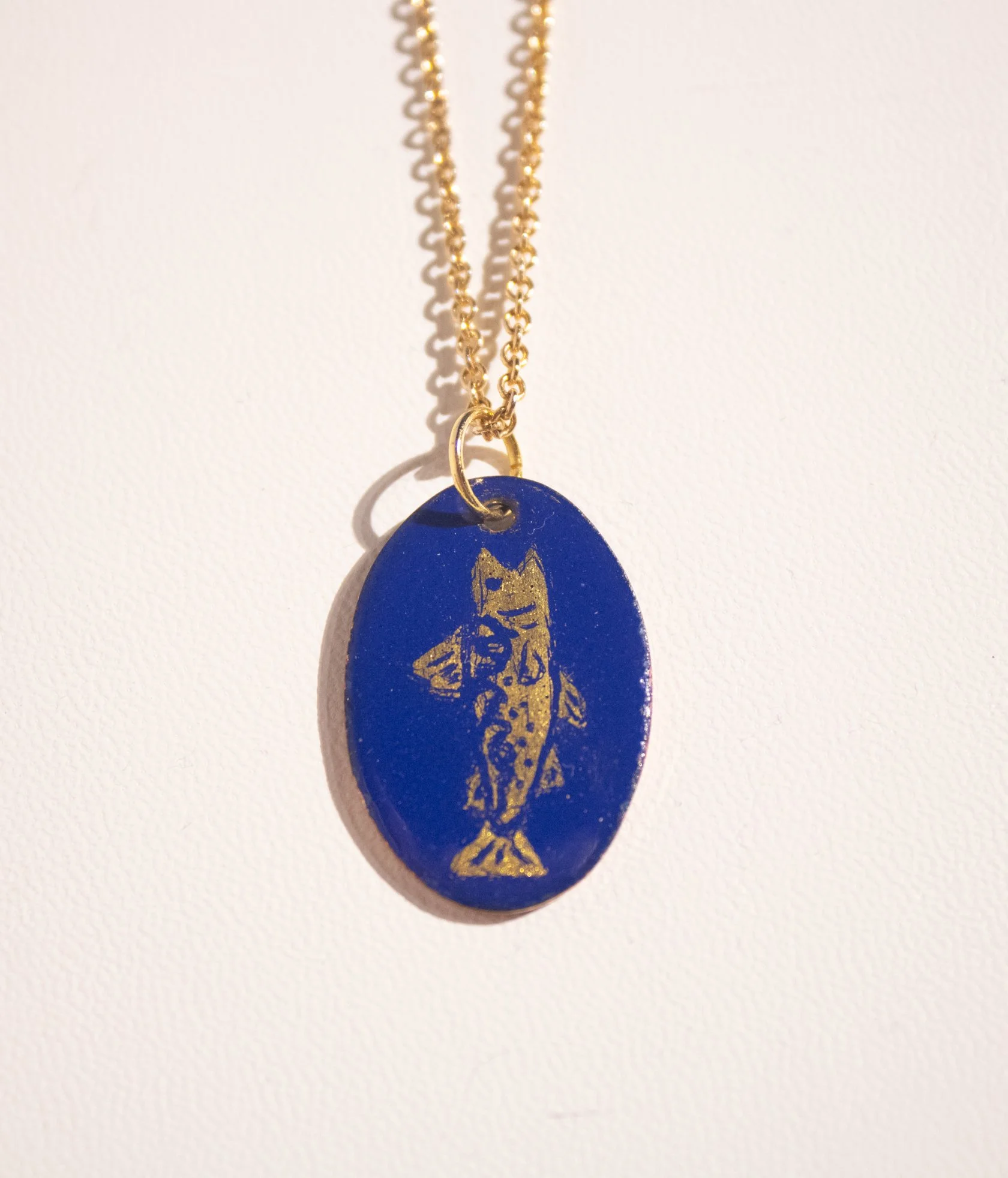 This small pendant features an oval shaped pendant enameled blue with a gold leaf brook trout figure depicted on it. 