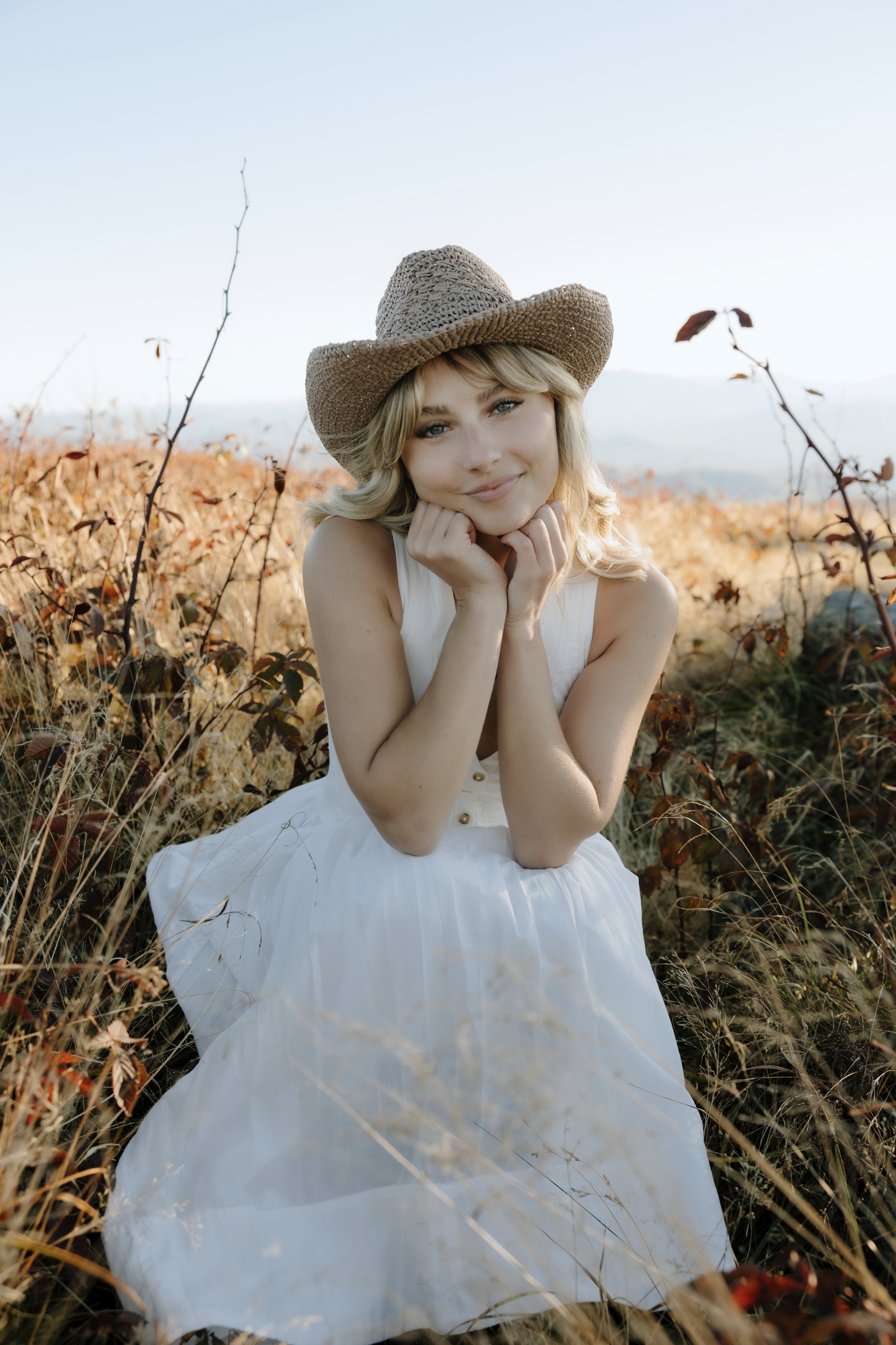 The artist, Haley, has short blonde hair and blue eyes. She Is wearing a white dress with buttons on the front and a wicker cowgirl hat. Her head is in her hands with her elbows propped on her knees in a sitting position, and she is smiling.
