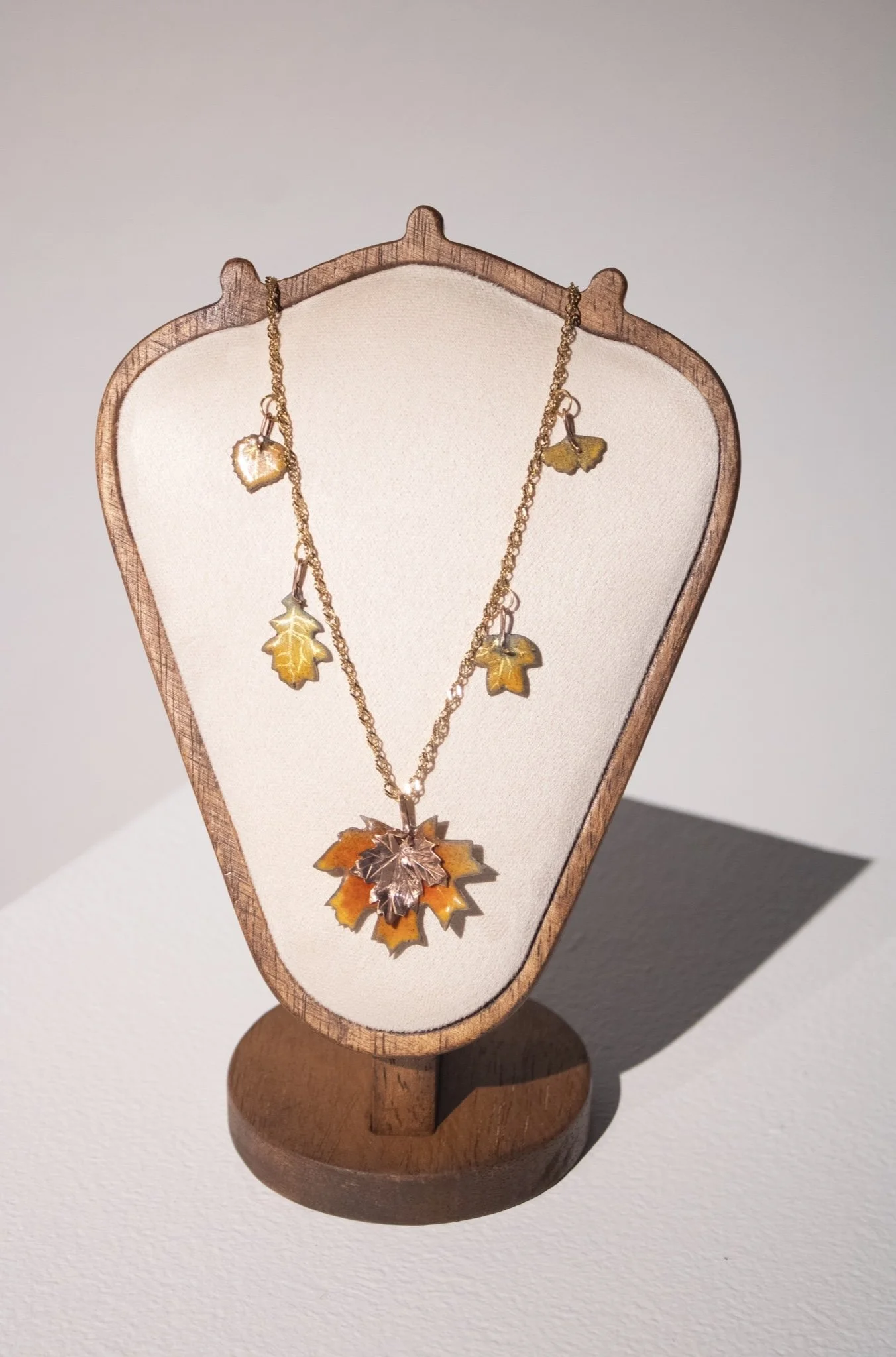 Titled, "Ode To Autumn", this gold charm necklace features five enameled leaf charms in mottled shades of orange, green, yellow, and red, with gold leaf sgraffito veins. The largest charm is a layered maple leaf at the center of the pendant - the bot
