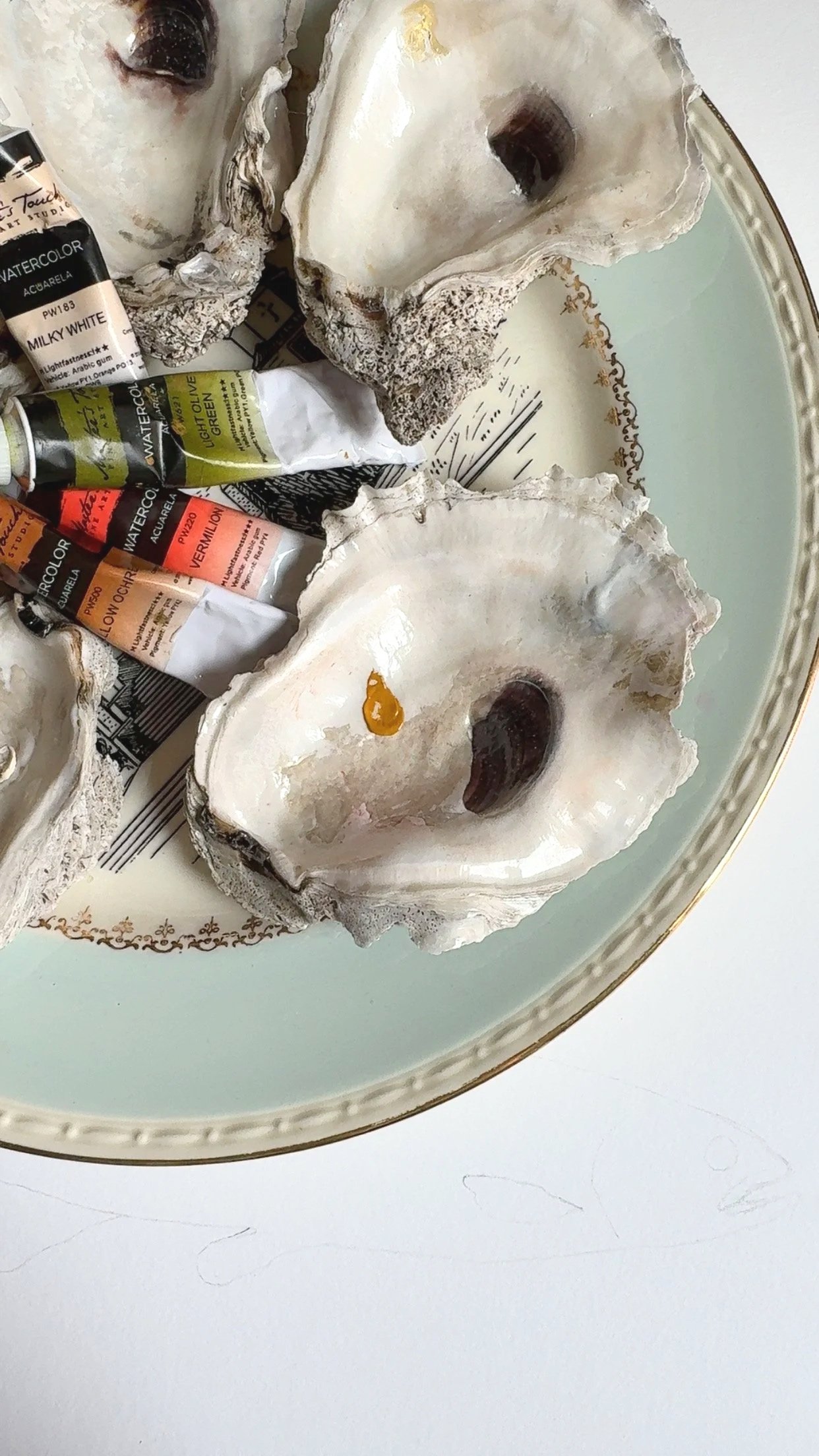 A closeup of a paint palette constructed from a fine china plate and oyster shells. Tubes of colorful paint lay on it and a dab of yellow paint sits in one of the oyster shells, awaiting use.