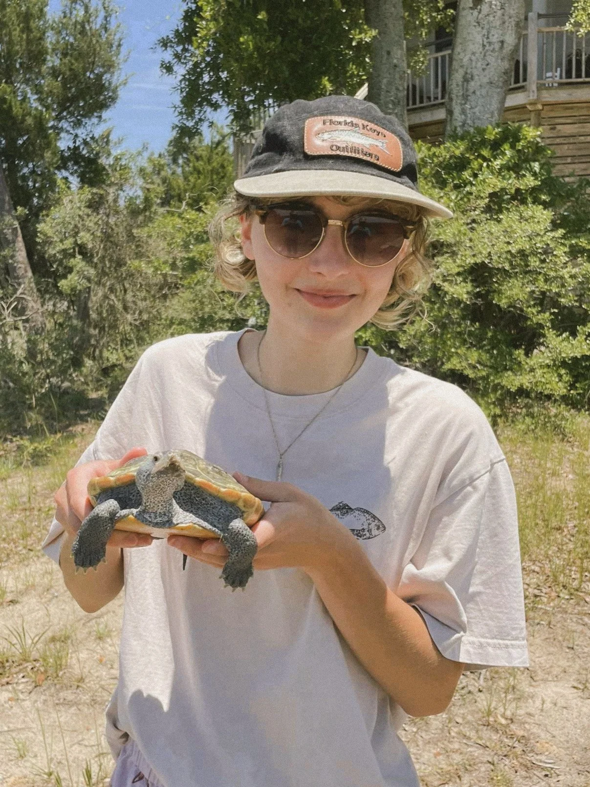 Haley wears sunglasses and a Florida Keys Outfitters hat brandishing a tarpon fish while she stands in the sun holding a small freshwater turtle in her hands. She is visiting a friend in South Carolina