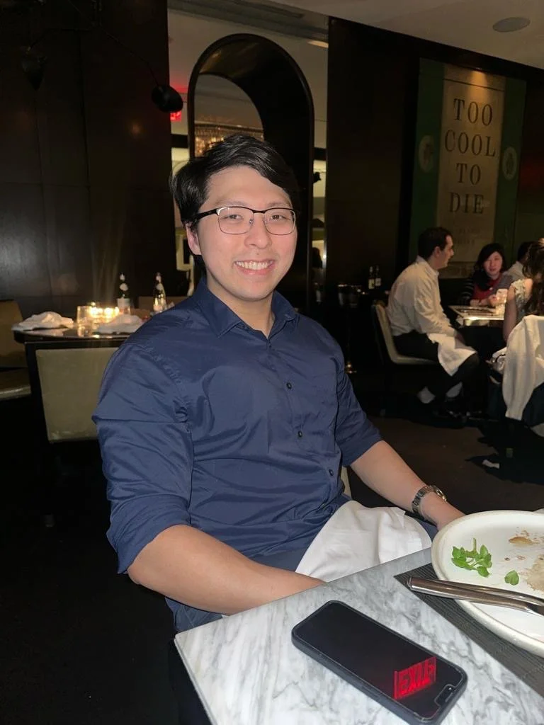 A smiling man wearing glasses and a blue shirt sitting at a restaurant table with an empty plate, a phone displaying a red glow, and a wristwatch. The restaurant has dark walls and other diners in the background.