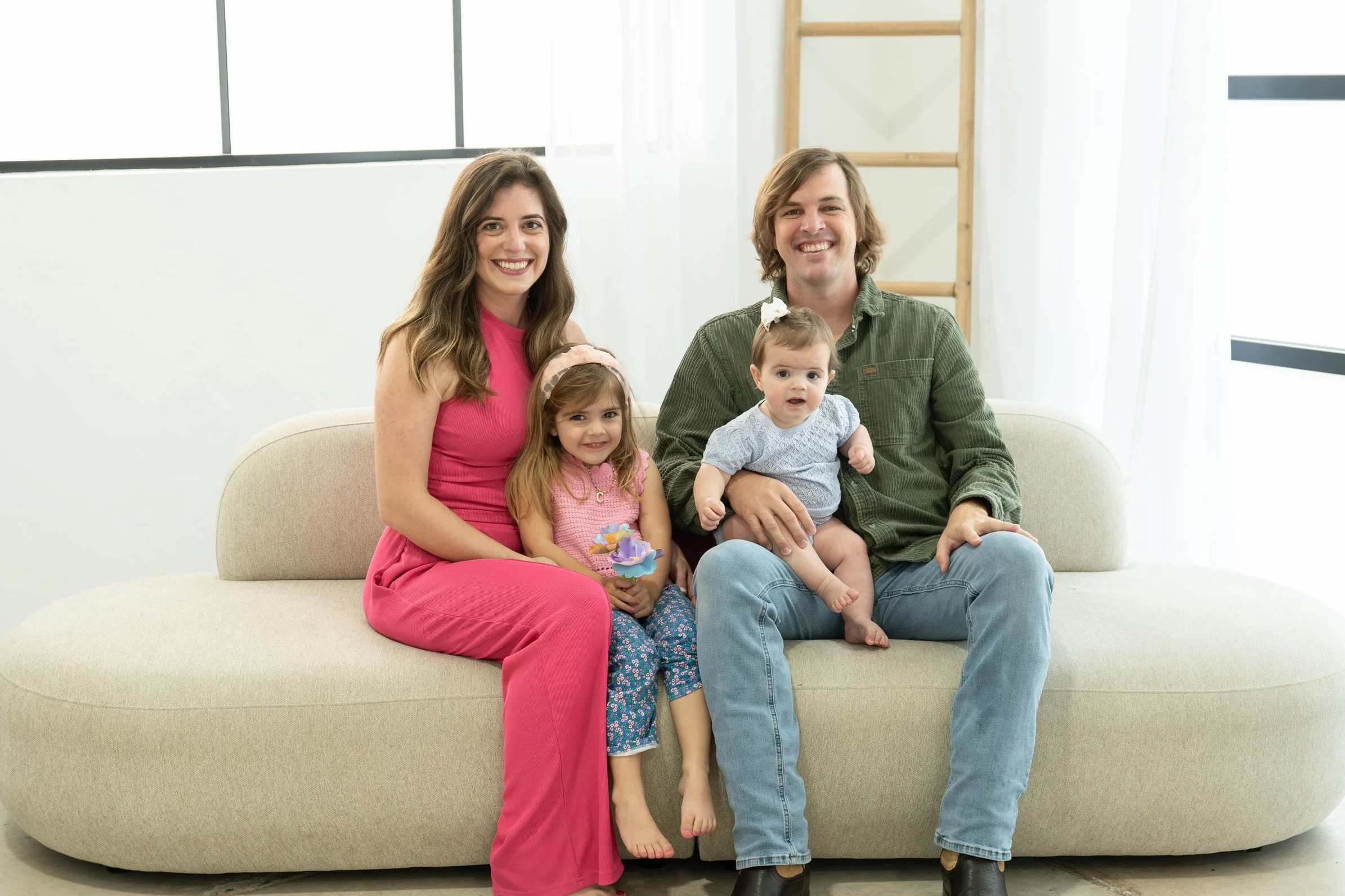 A smiling family of four sitting on a beige sofa in a bright living room. The mother, wearing a pink sleeveless dress, and the father, dressed in a green shirt and jeans, are seated with two young girls, one sitting on the mother's lap and the other sitting on the father's lap. The two girls are dressed in colorful clothes, with the older girl holding a small toy.