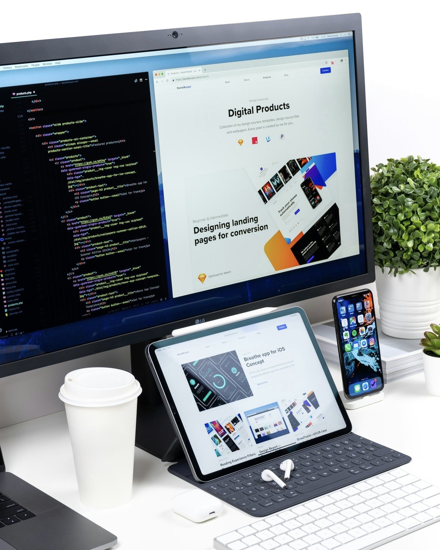 A computer monitor displaying coding on the left and a website for digital products on the right, with a tablet, smartphone, white coffee cup, keyboard, wireless earphones, and a potted plant on a white desk.