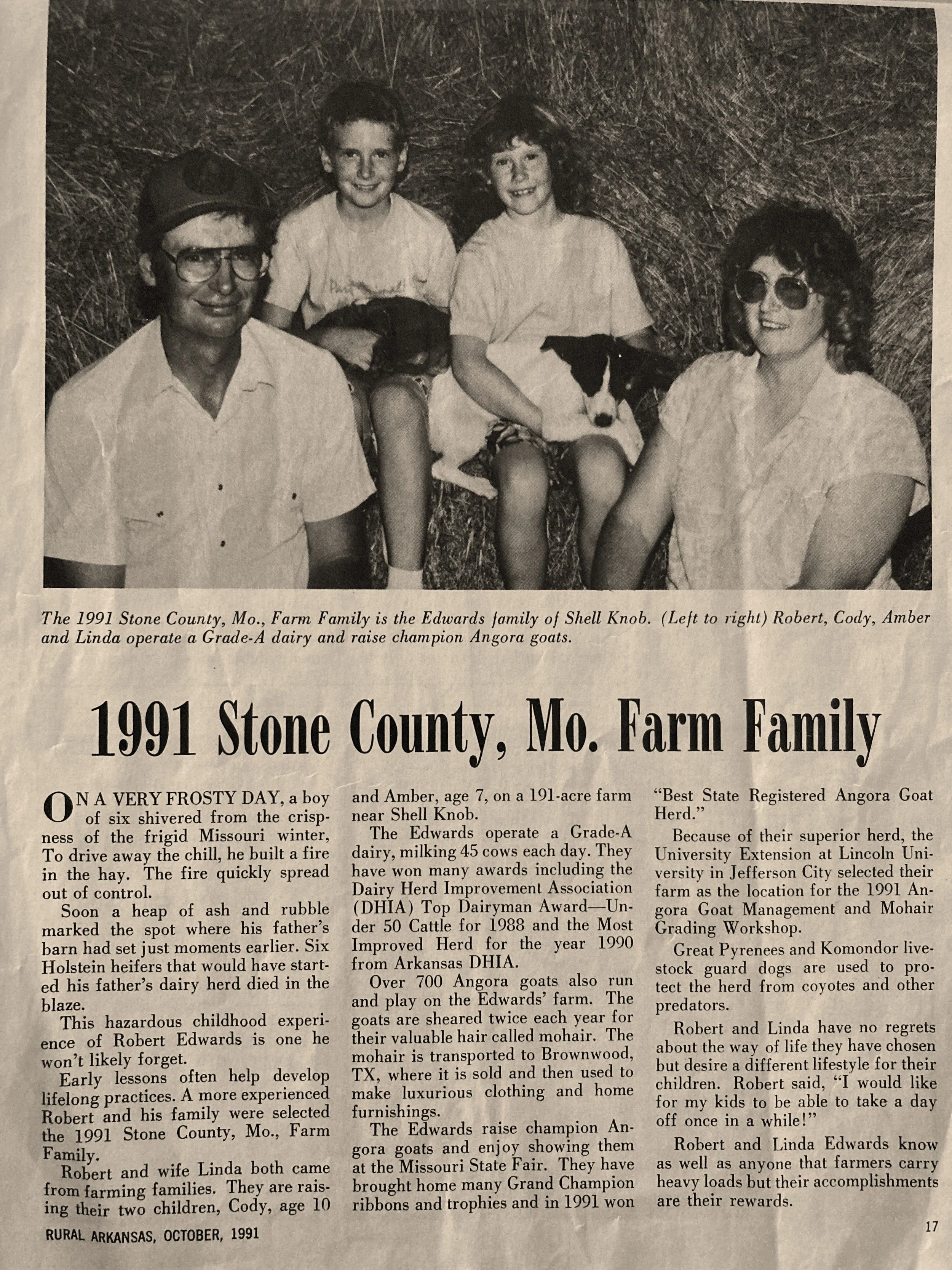 Black and white photo of a family sitting outdoors on a farm, with two children, two adults, and a dog, all smiling. The father is wearing glasses and a hat, and the mother is wearing sunglasses.