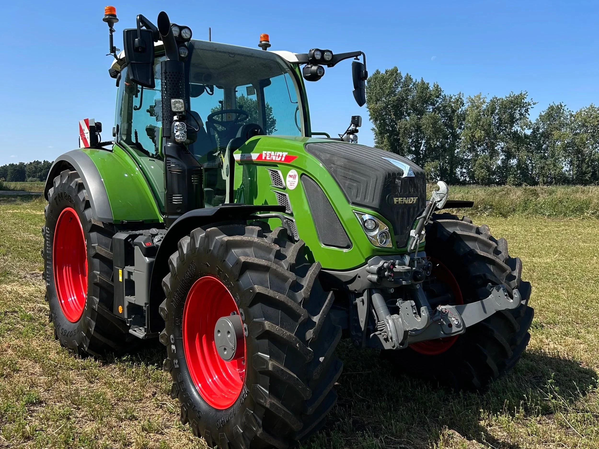 A green modern tractor with red wheels parked on a grassy field under a blue sky with trees in the background.