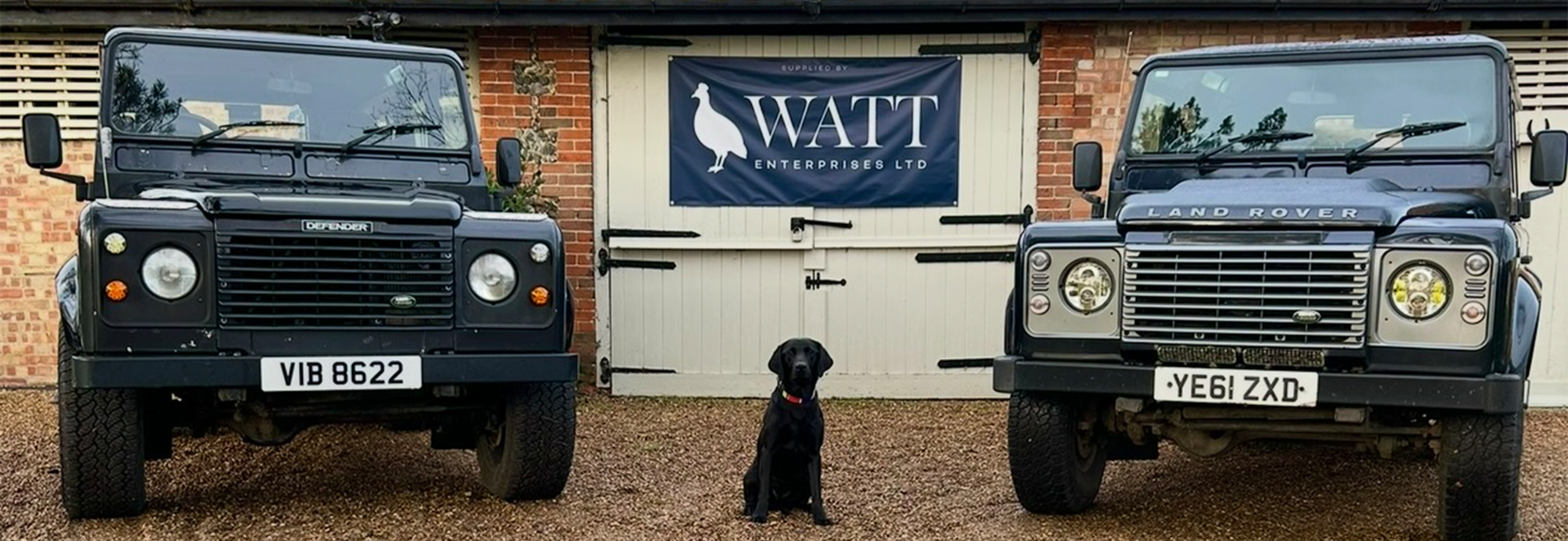 Two black Land Rover Defender vehicles parked in front of a white gate with a "WATT Enterprises Ltd" banner, and a black dog sitting between them.