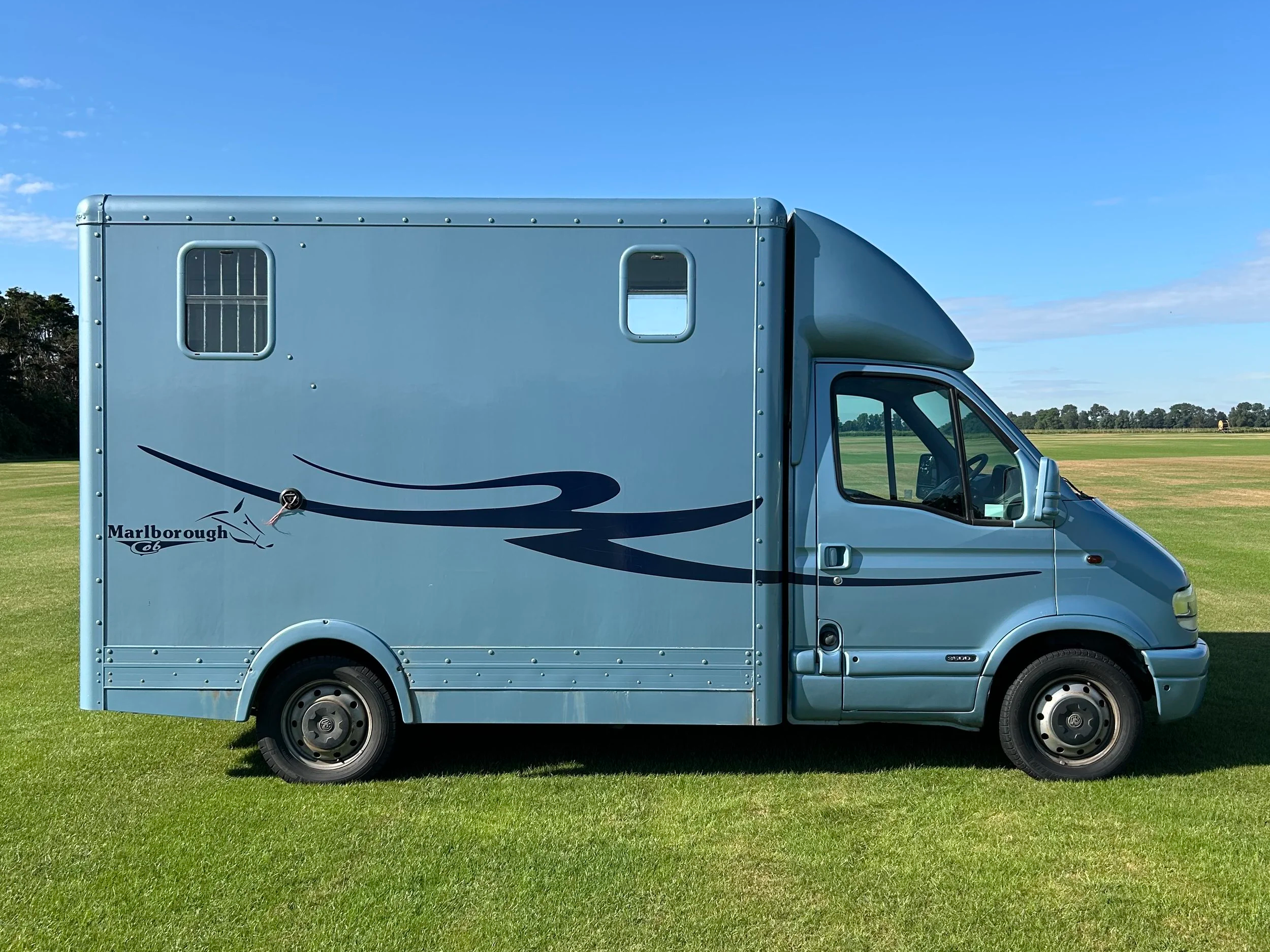 Blue delivery truck with a stylized swoosh logo and the word 'Marlborough' on the side, parked on a grassy field with a blue sky and trees in the background.