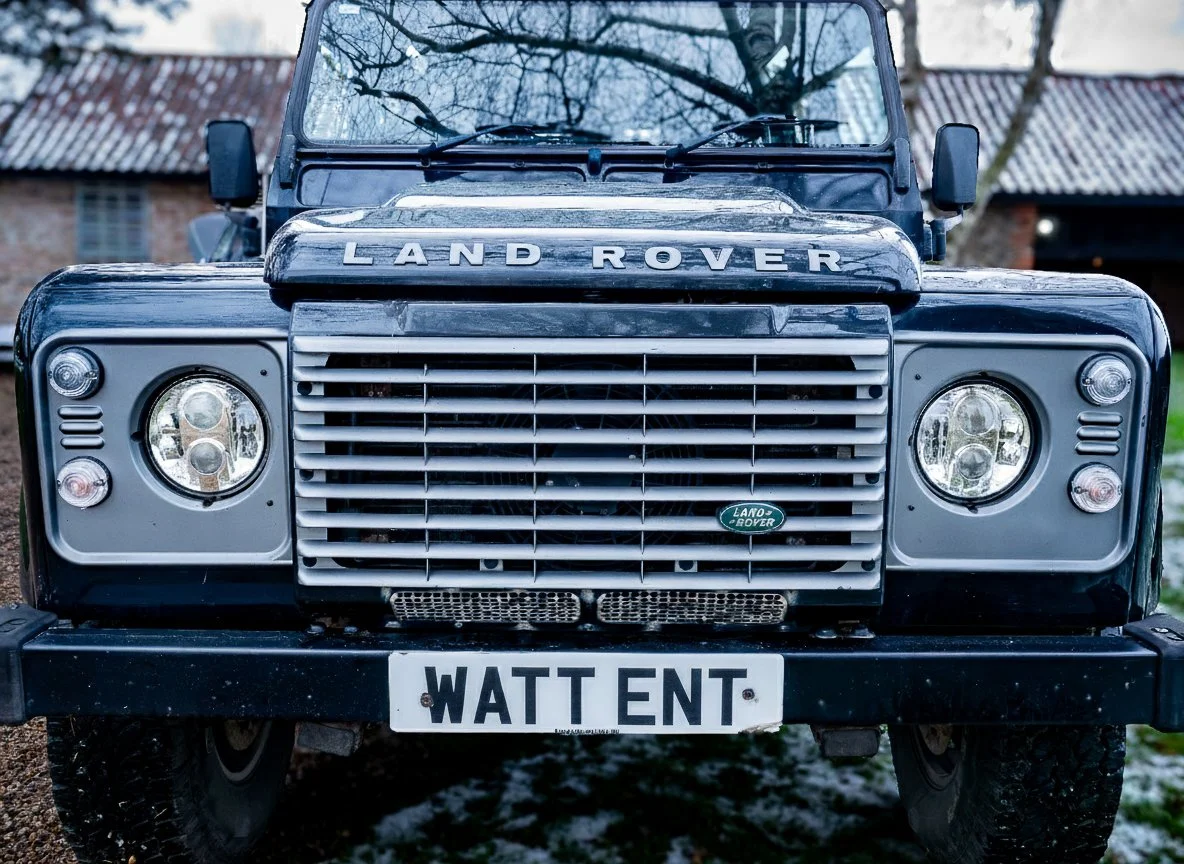Front view of a black Land Rover SUV with a custom license plate reading 'WATT ENT,' parked outdoors near old brick buildings and leafless trees.