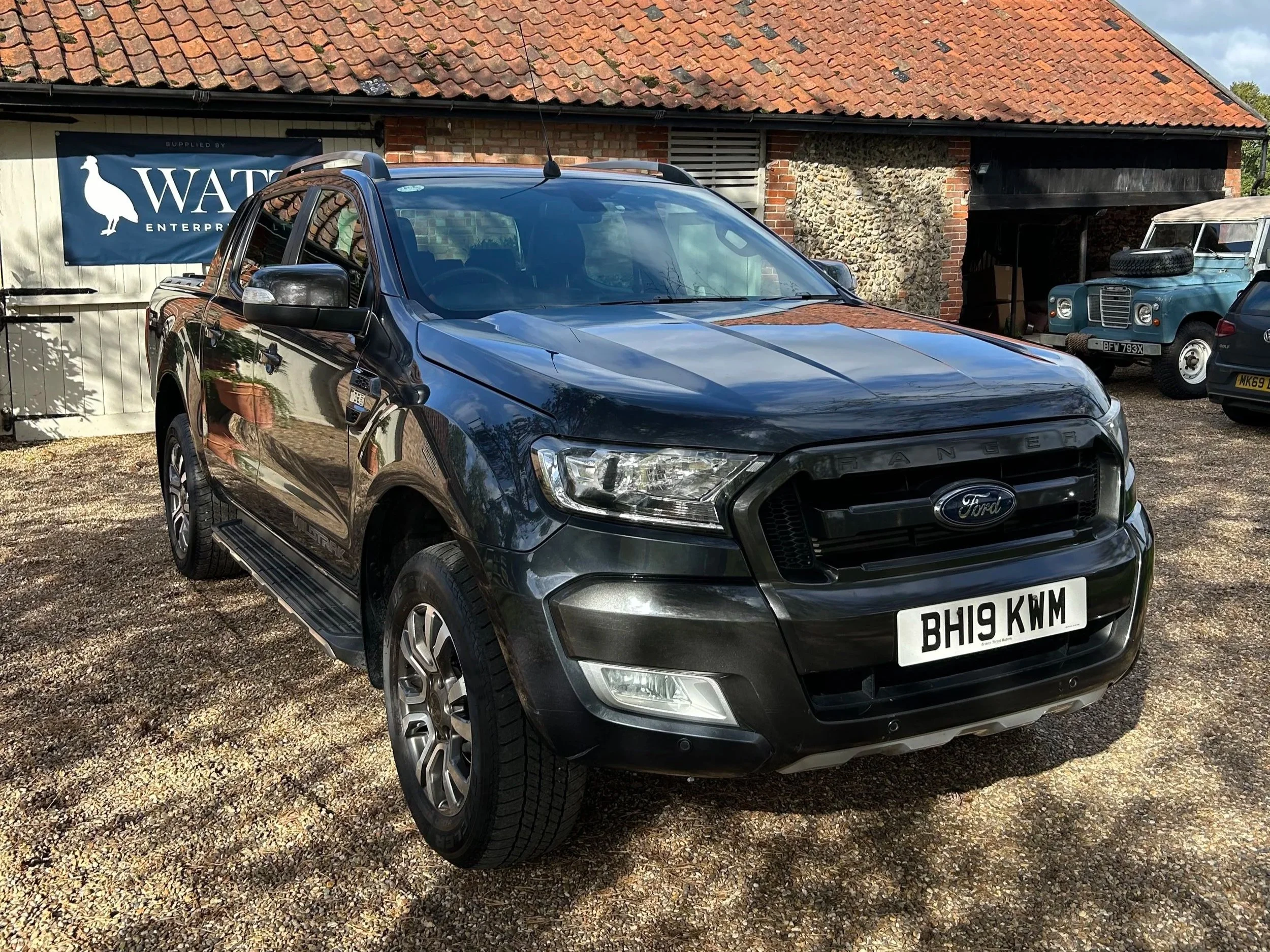 Black Ford Ranger pickup truck parked on gravel in front of a building with a banner that reads "Water Enterprises". Two vintage vehicles are parked nearby.