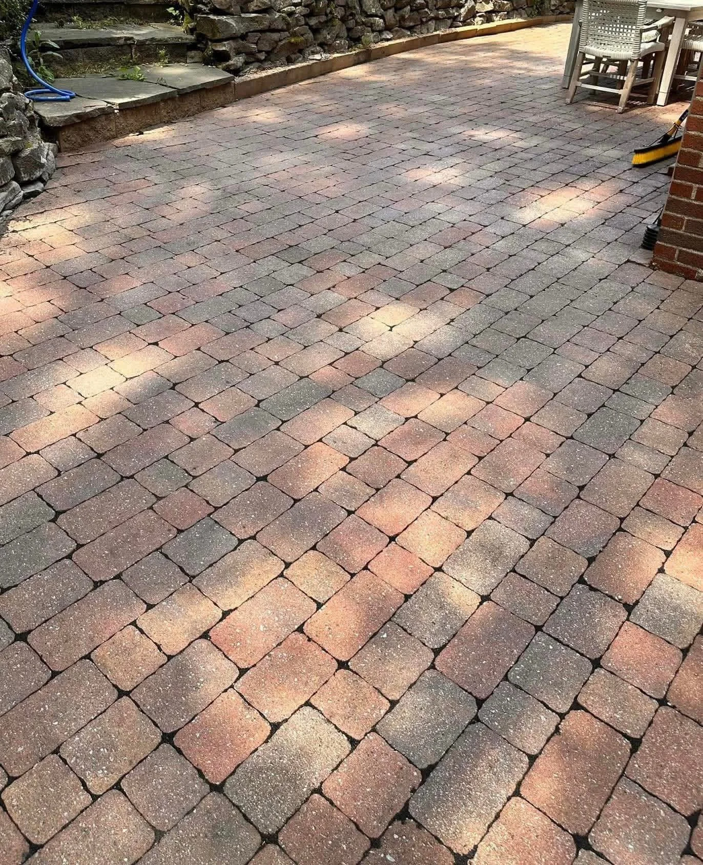 A paved outdoor patio area with brick pavers, surrounded by a stone wall on one side and a brick wall on the other, with outdoor furniture and some tools visible.