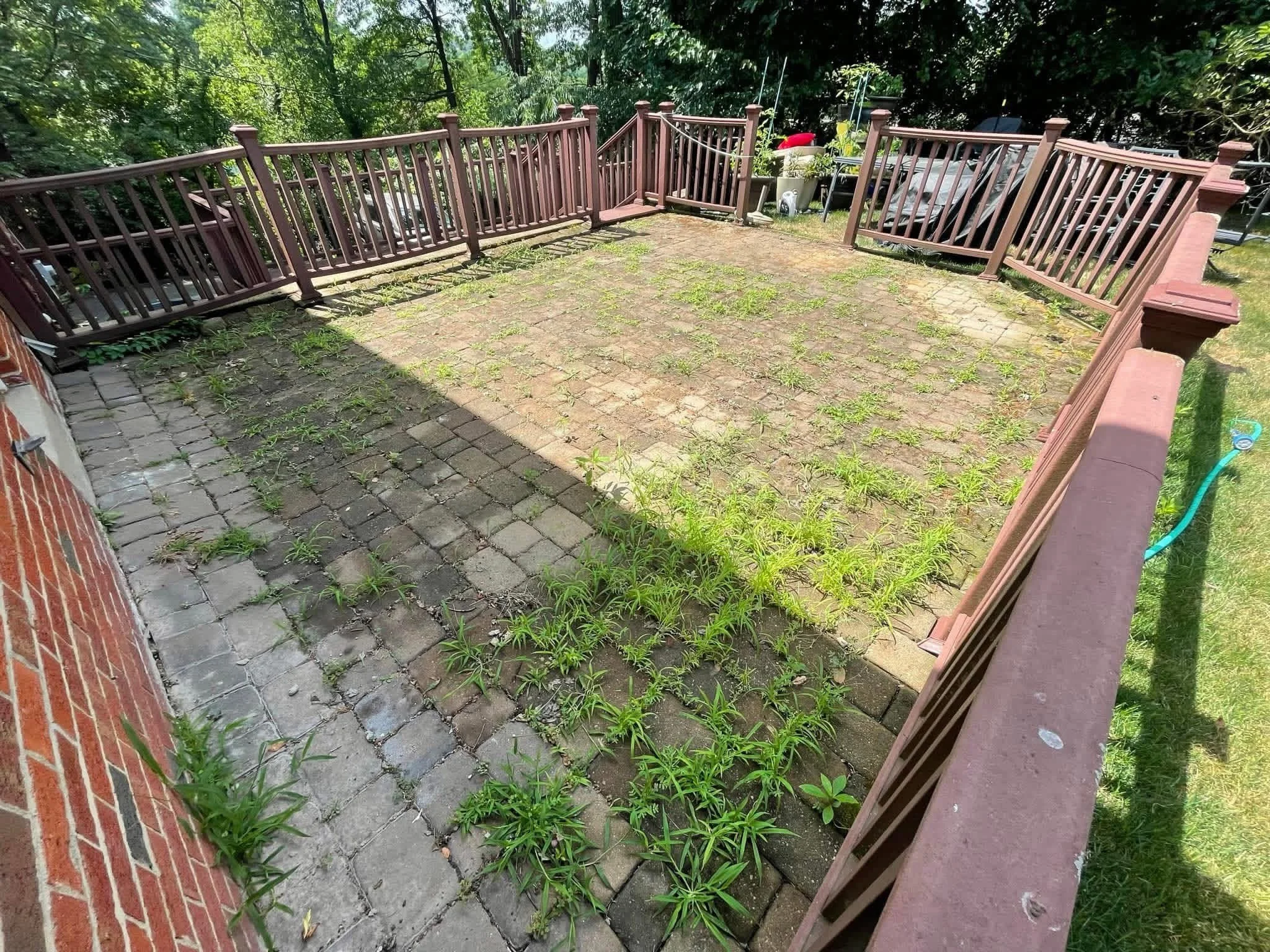 Backyard patio with brick and paver stones, surrounded by a wooden railing, with patches of grass and weeds growing between the stones, and trees in the background.