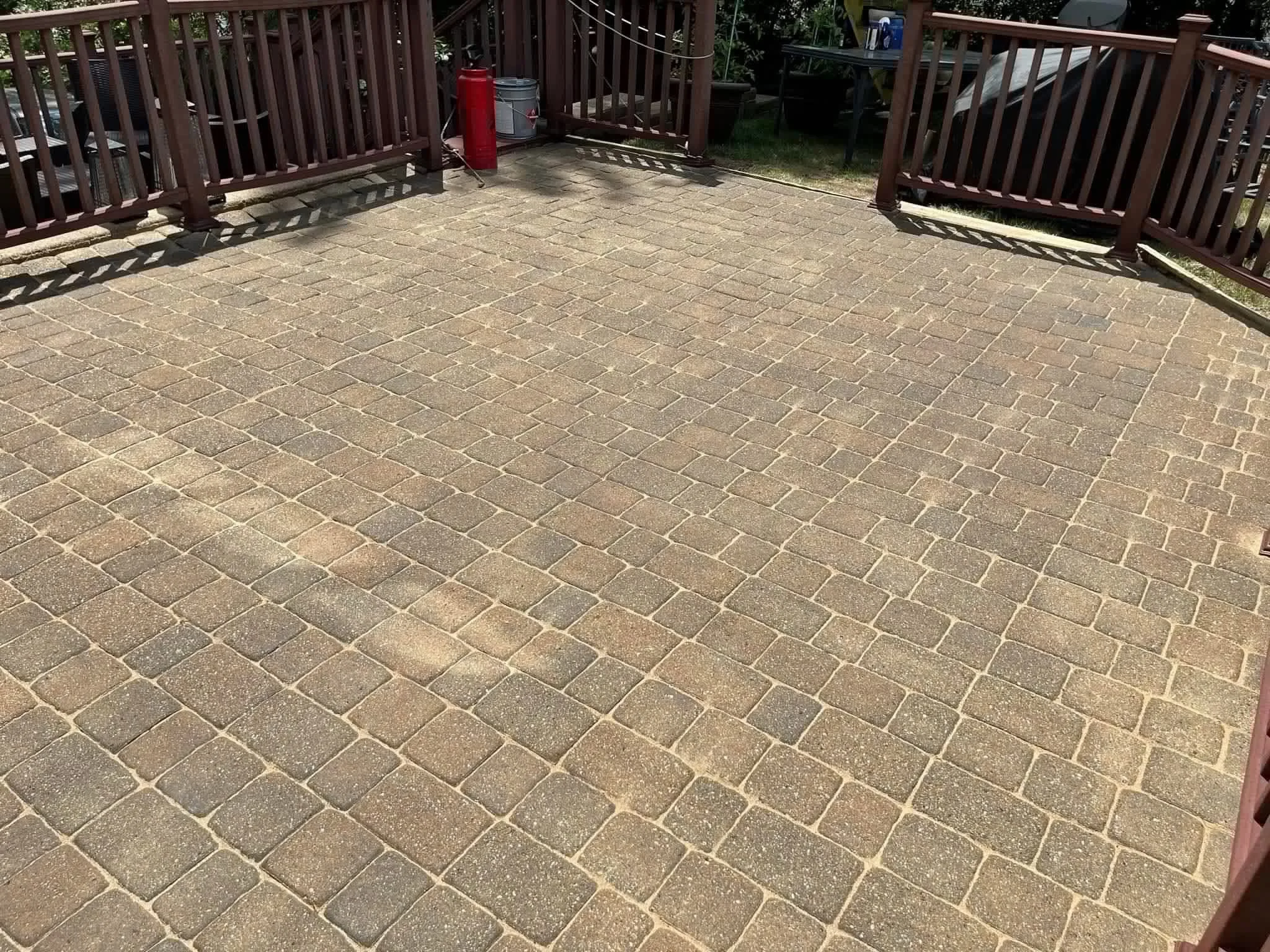 An outdoor patio area with brick paving stones, surrounded by wooden fence panels. There are some red and gray containers and a few trees and grass visible beyond the fence.