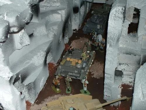 A narrow corridor of destruction—armor advances single file while infantry scan the upper floors for movement.