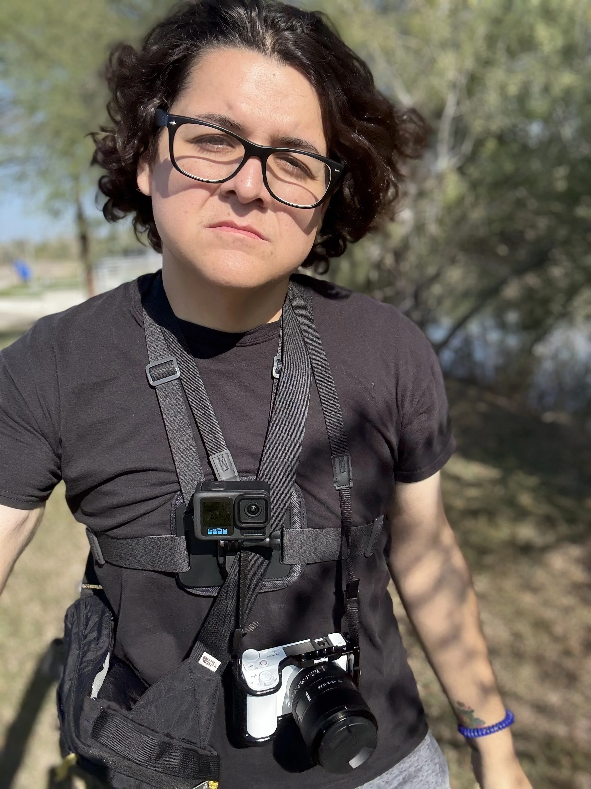 A woman with curly dark hair, glasses, and tattoos on her left arm, wearing a black t-shirt, outdoors on a sunny day with trees and a lake in the background. She has a camera hanging around her neck and a GoPro mounted on her chest strap.