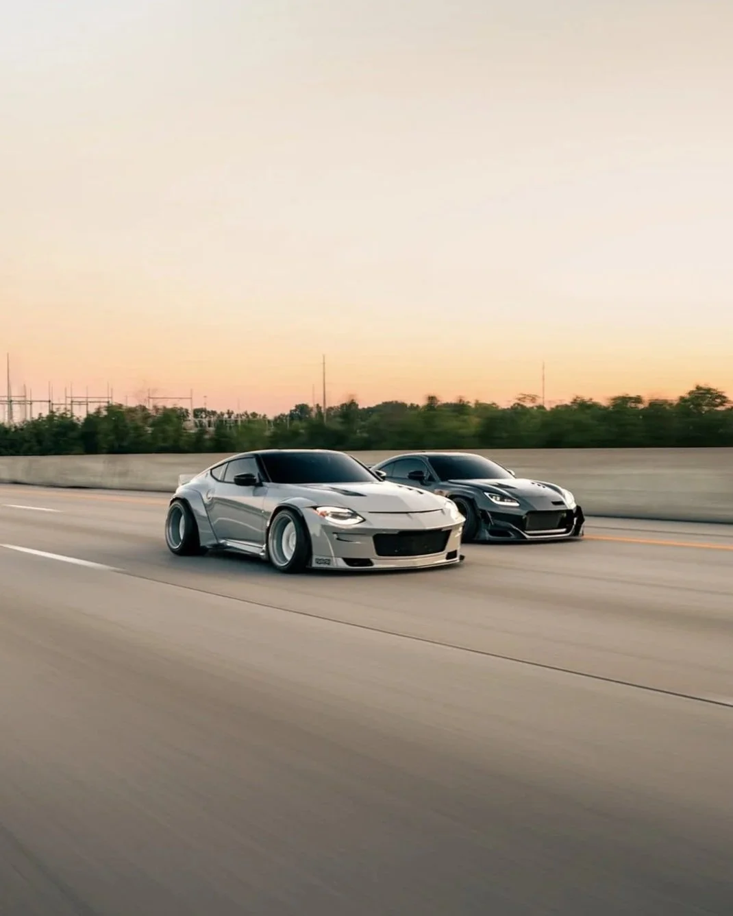 Two modified sports cars, one silver and one black, racing on a highway during sunset.