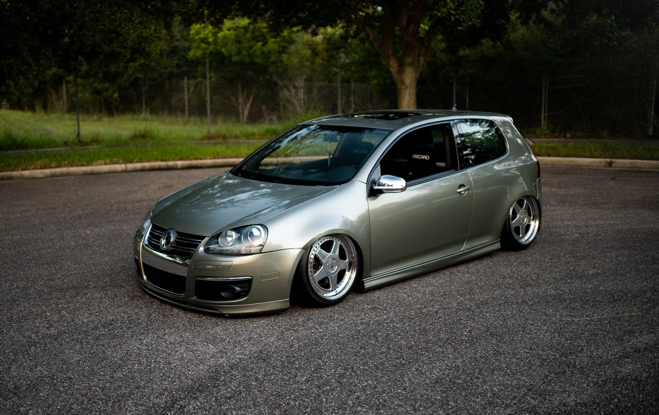A lowered silver Volkswagen Golf hatchback parked on a paved road with green grass and trees in the background.
