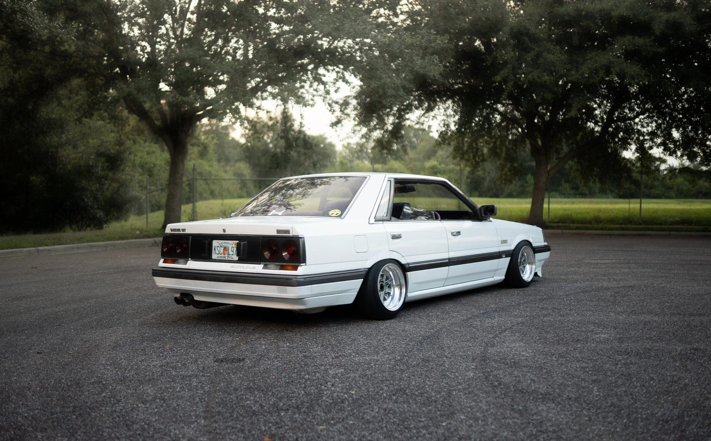 White vintage Nissan car parked on a paved lot with trees and a grassy field in the background.