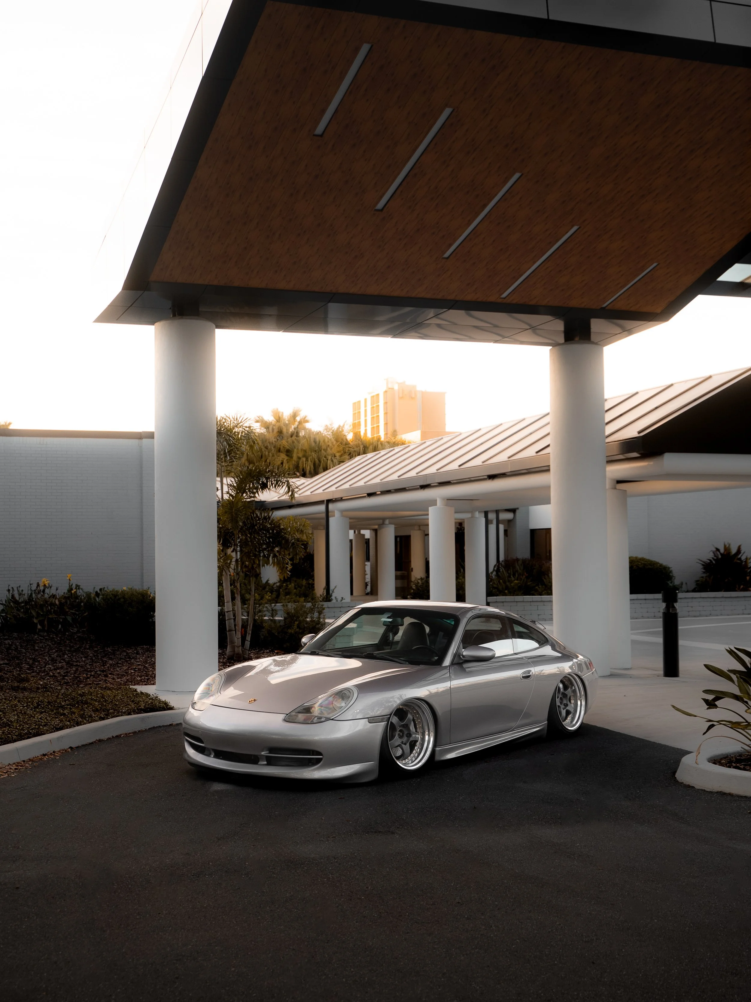 Silver Porsche sports car parked under a modern building with white columns and a wooden ceiling, in an urban setting during sunset.