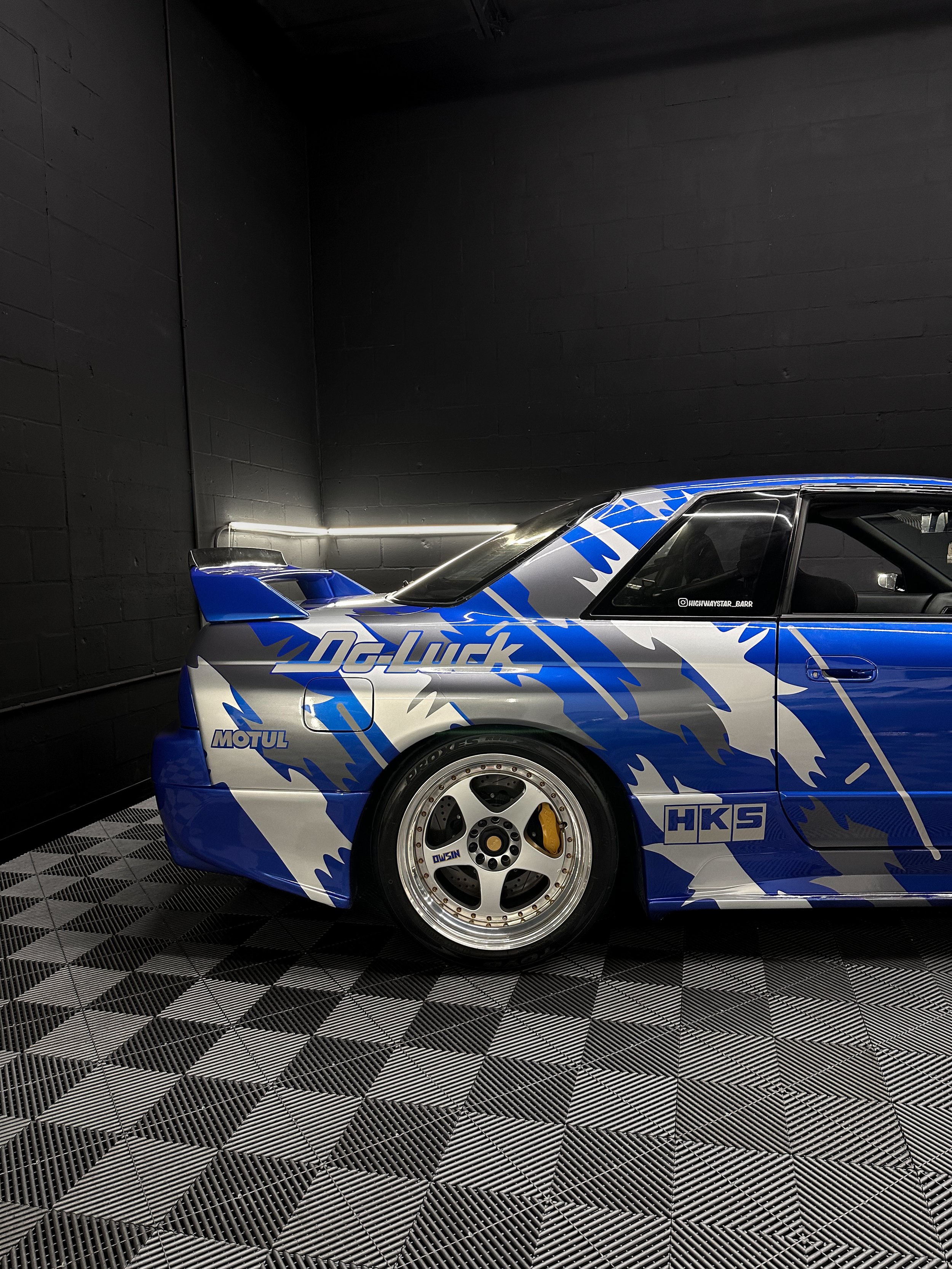 A blue and silver race car with sponsor logos, parked on a checkered floor in a black-walled room.