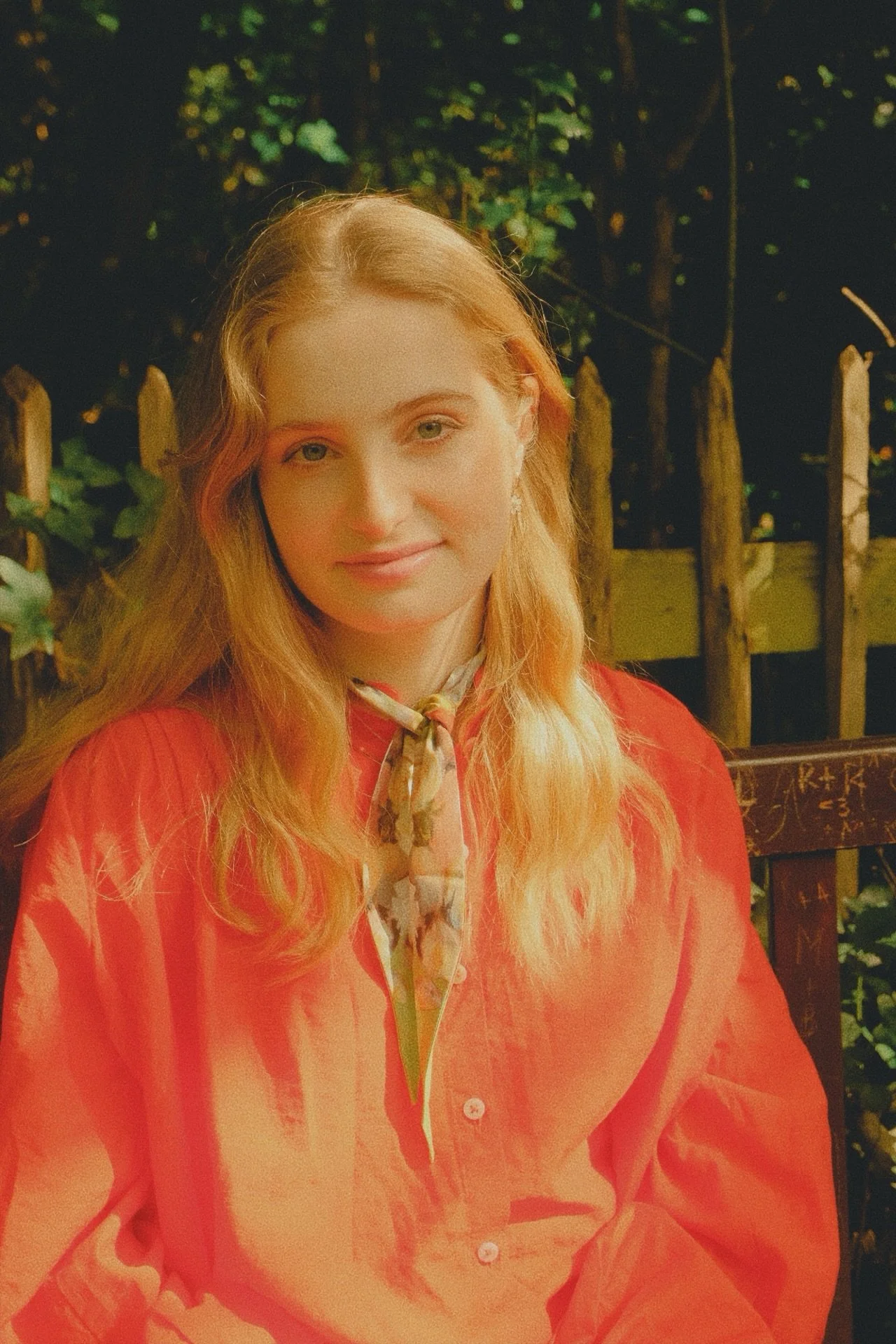 A woman with long, wavy, blonde hair wearing a coral-colored shirt and a patterned scarf around her neck, sitting outdoors with a wooden fence and greenery in the background.