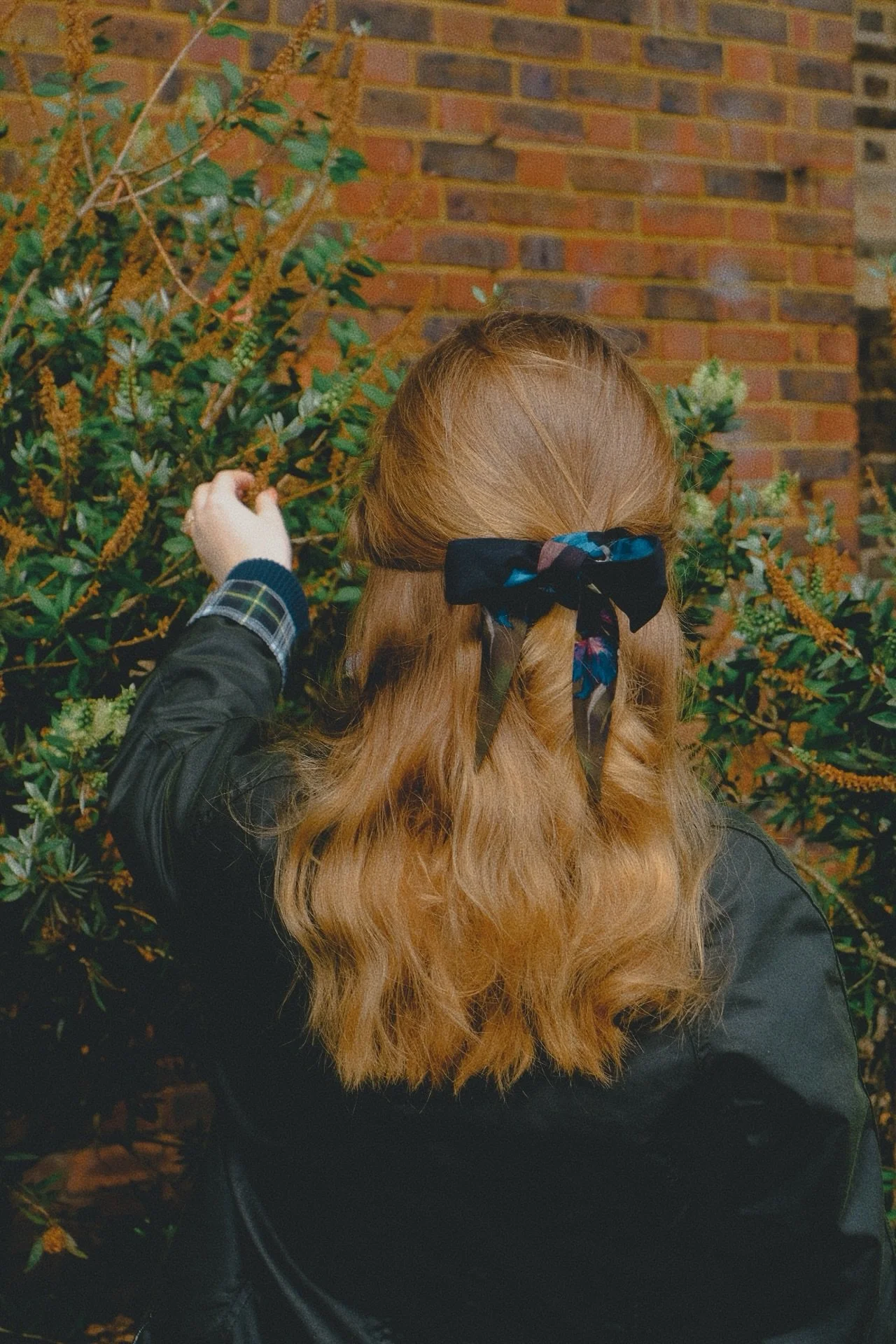Back view of a woman with long wavy red hair tied with a black bandana, reaching out to touch a green leafy plant with yellow flowers, against a brick wall background.