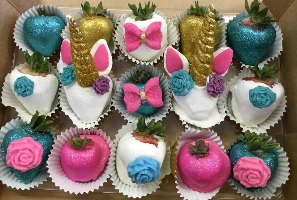 A box of decorated strawberries with various colors and designs, including pink, blue, white, and gold accents, some with bows and roses, and others with glitter and gold sprinkles.
