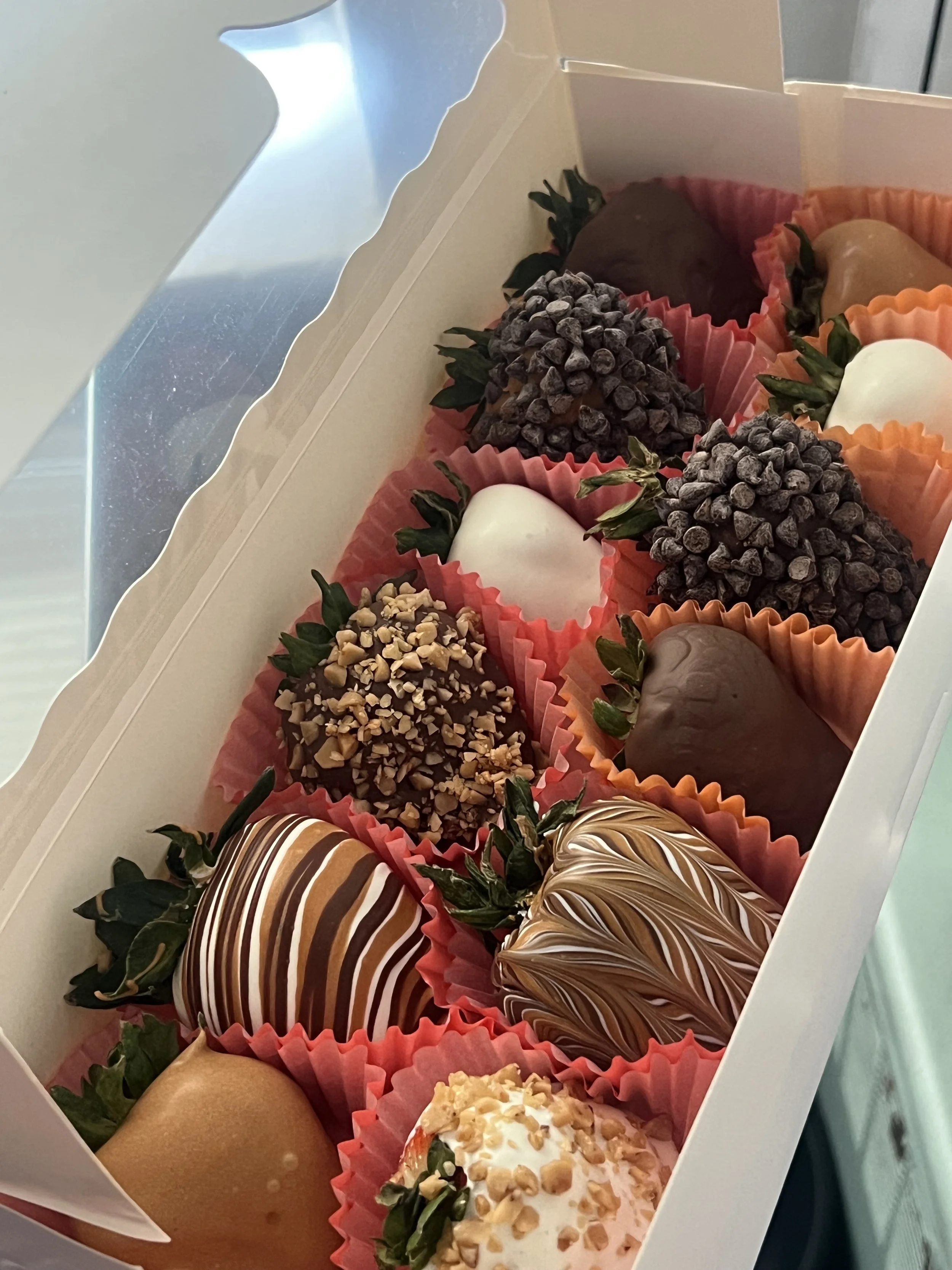 A box of assorted chocolate-covered strawberries decorated with various toppings such as nuts, chocolate chips, and drizzles, each placed in pink cupcake liners.