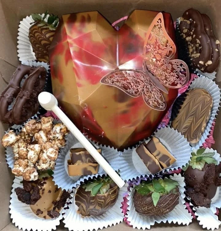 A heart-shaped cake with a butterfly decoration on top, surrounded by various chocolates and candies in paper cups.