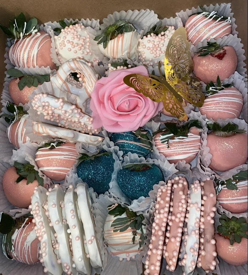 Decorative assortment of strawberries dipped in pink and white coatings, some with sprinkles and drizzle, arranged with a pink rose, a gold butterfly, and glittery blue spheres.